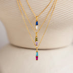 The Toi et Moi Tapered Birthstone Necklace, featuring a stunning baguette-cut pendant and colorful charms, is displayed on a cream textured stand alongside other gold necklaces.
