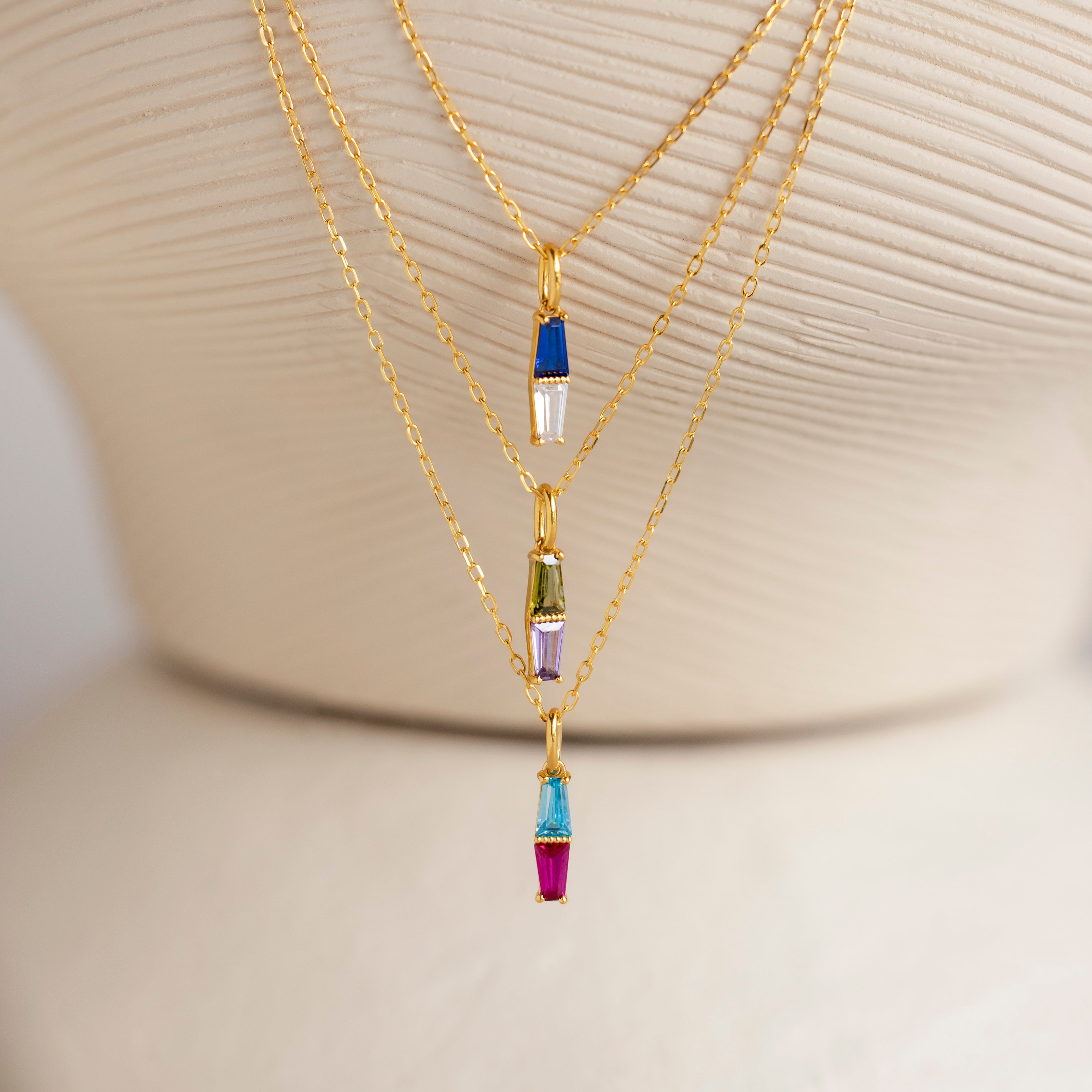 The Toi et Moi Tapered Birthstone Necklace, featuring a stunning baguette-cut pendant and colorful charms, is displayed on a cream textured stand alongside other gold necklaces.