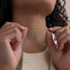 A woman holds the Toi et Moi Tapered Birthstone Necklace, a delicate gold piece with a small pendant, close to her neck.