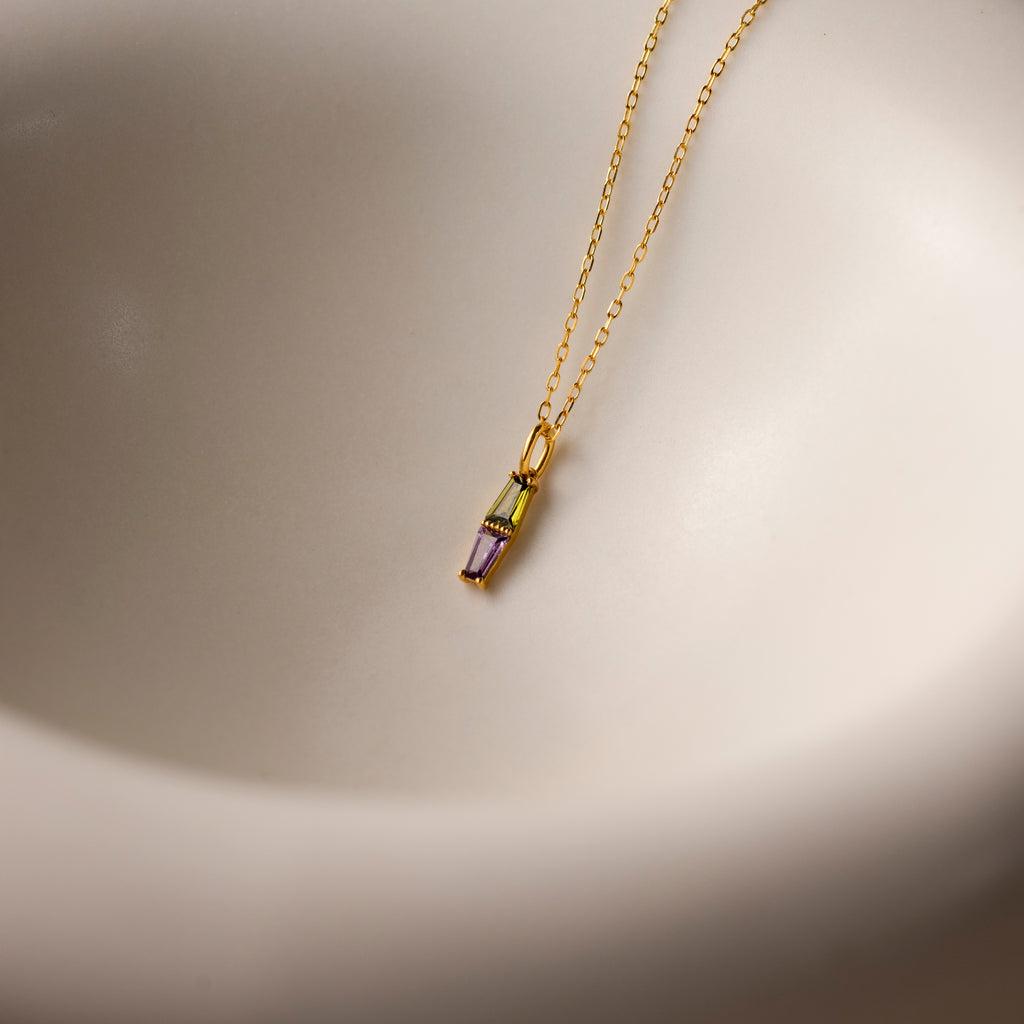 The Toi et Moi Tapered Birthstone Necklace showcases a small, baguette-cut pendant elegantly displayed on a smooth, white curved surface.