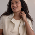 Smiling woman with curly hair wears a beige blazer, cream top, and the Toi et Moi Tapered Birthstone Necklace while sitting indoors.