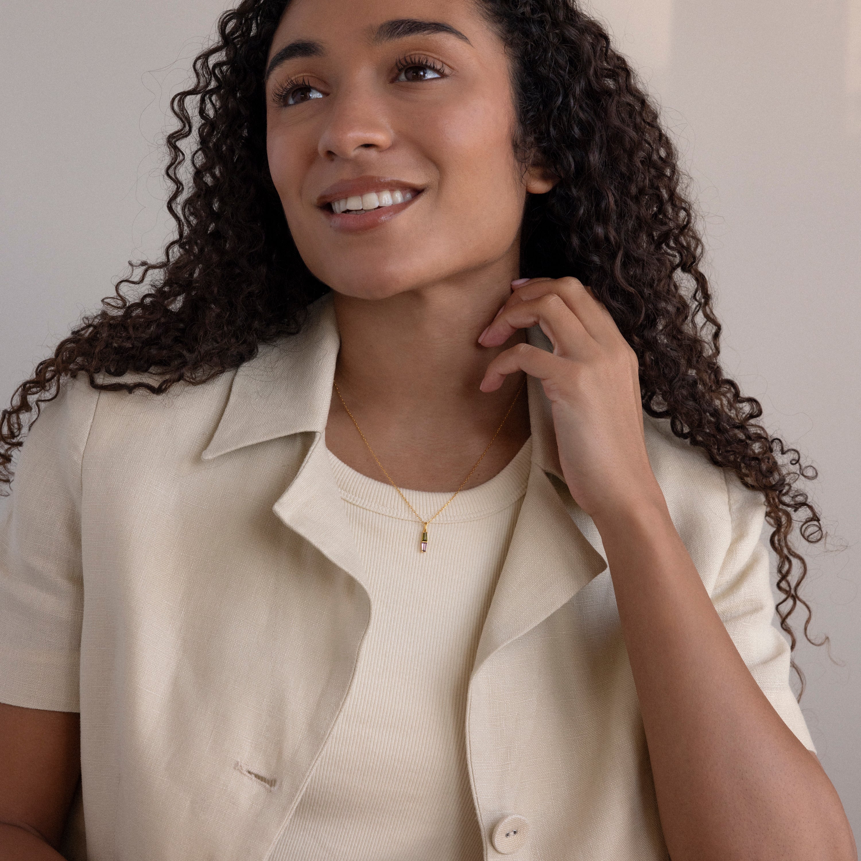 Smiling woman with curly hair wears a beige blazer, cream top, and the Toi et Moi Tapered Birthstone Necklace while sitting indoors.