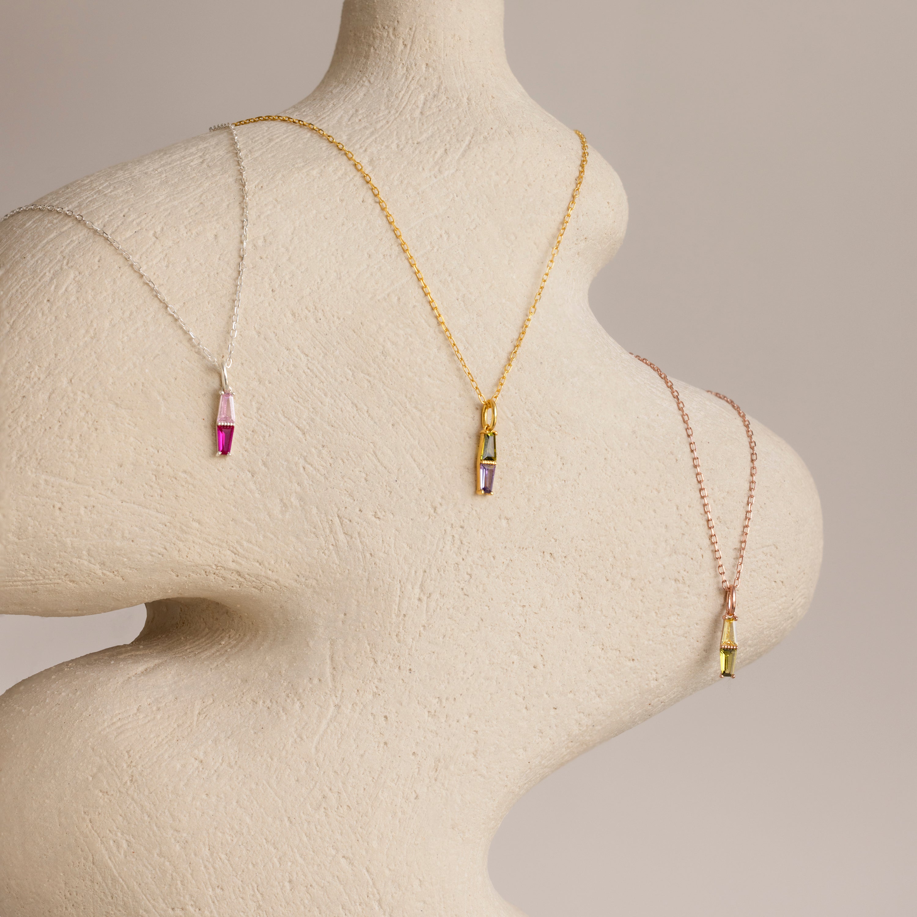 The Toi et Moi Tapered Birthstone Necklace, a dazzling colorful gemstone pendant, is elegantly draped on a textured, abstract mannequin torso.