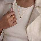 A person wearing gold rings and the Toi et Moi Tapered Birthstone Necklace, paired with a cream ribbed shirt and beige blazer.