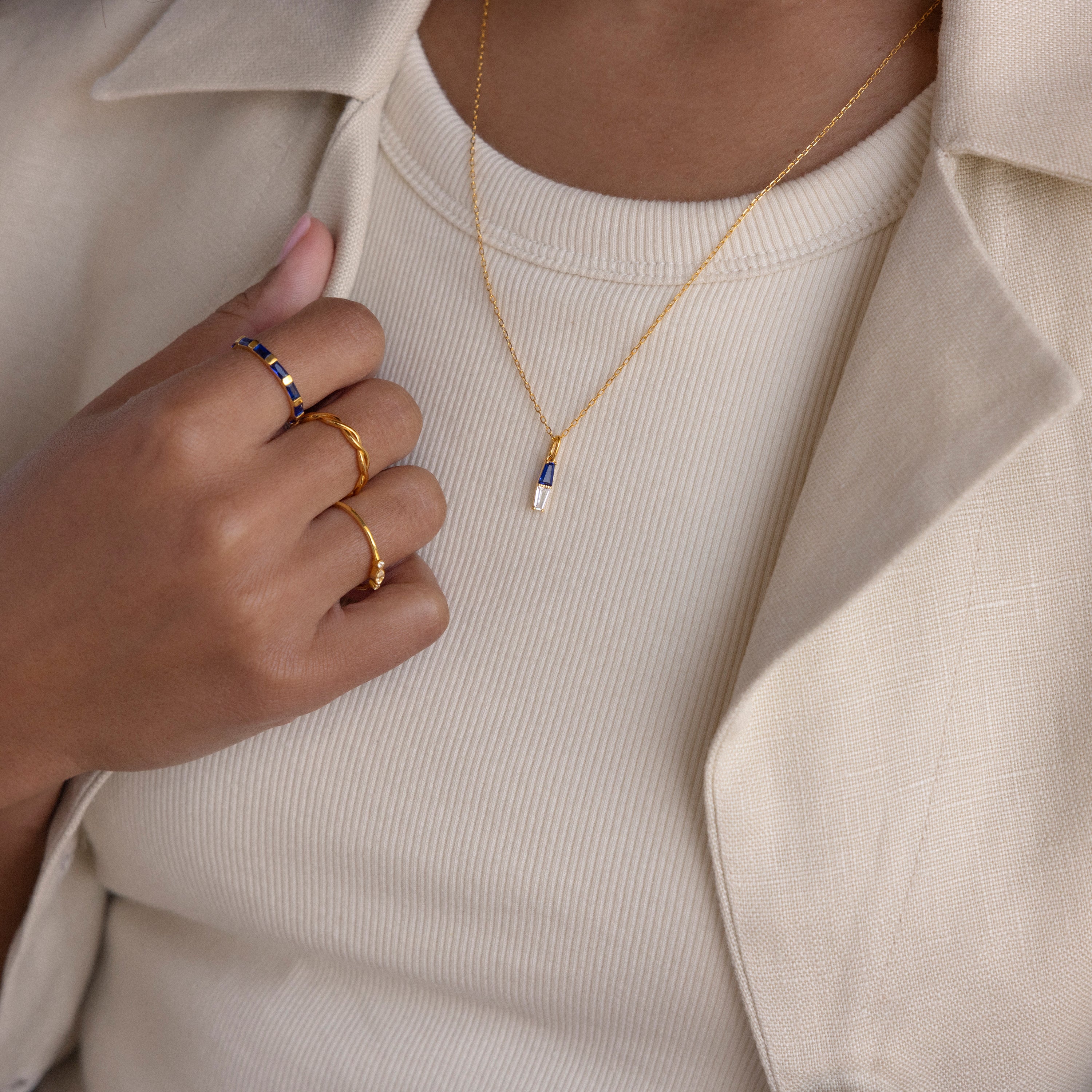 A person wearing gold rings and the Toi et Moi Tapered Birthstone Necklace, paired with a cream ribbed shirt and beige blazer.