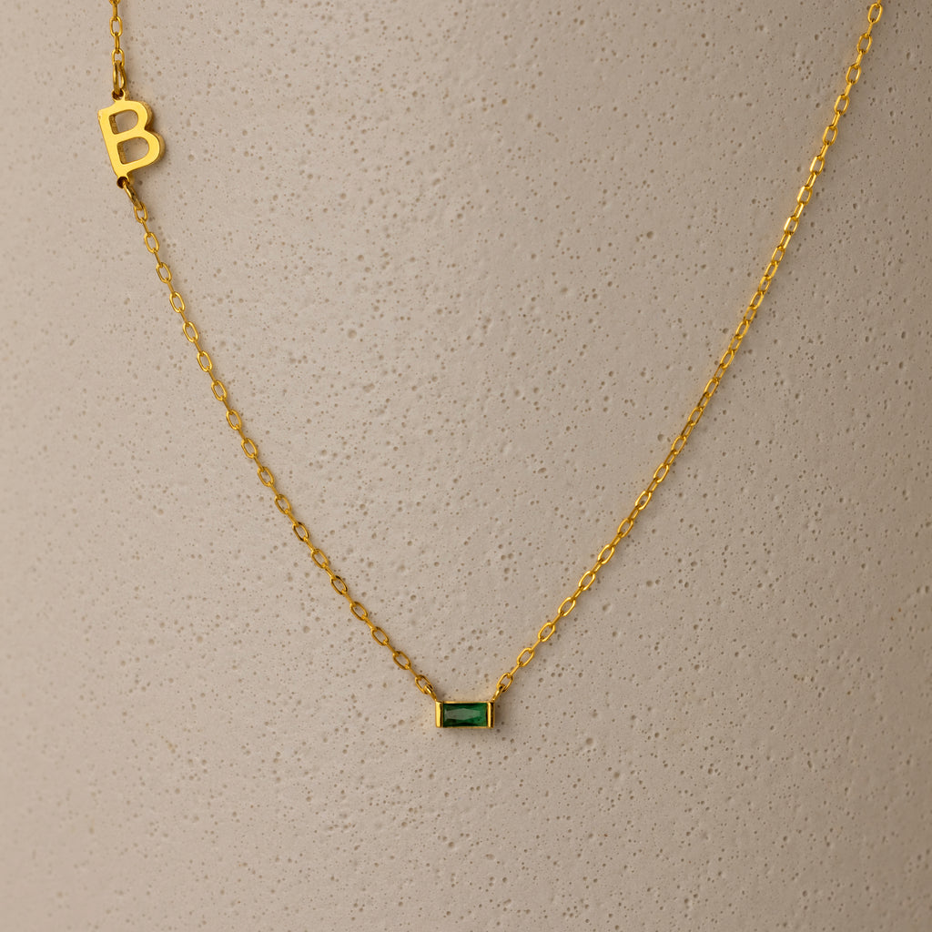 Initial Baguette Birthstone Necklace featuring a sideways initial charm and a baguette-cut birthstone pendant, shown on a textured beige surface.