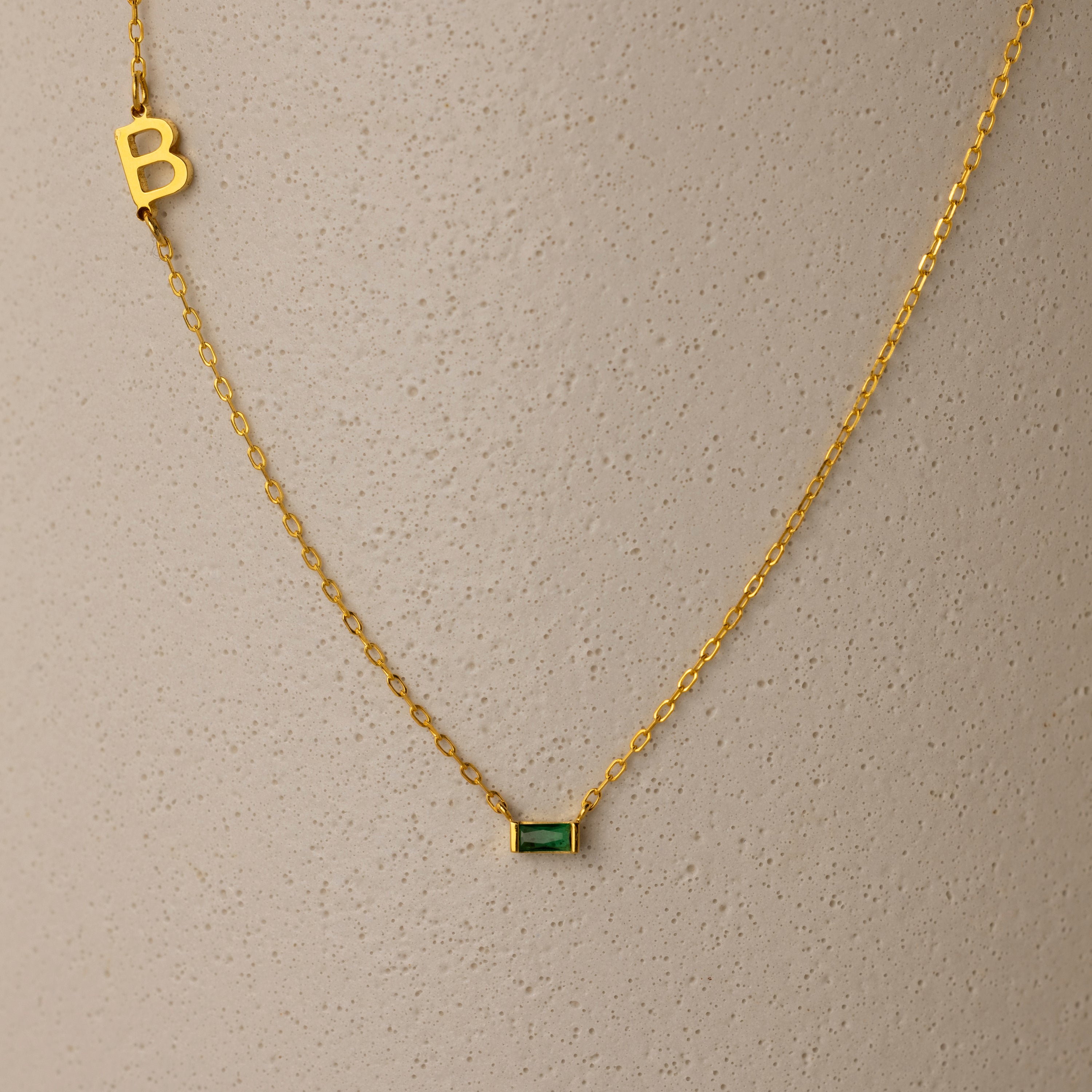 Initial Baguette Birthstone Necklace featuring a sideways initial charm and a baguette-cut birthstone pendant, shown on a textured beige surface.
