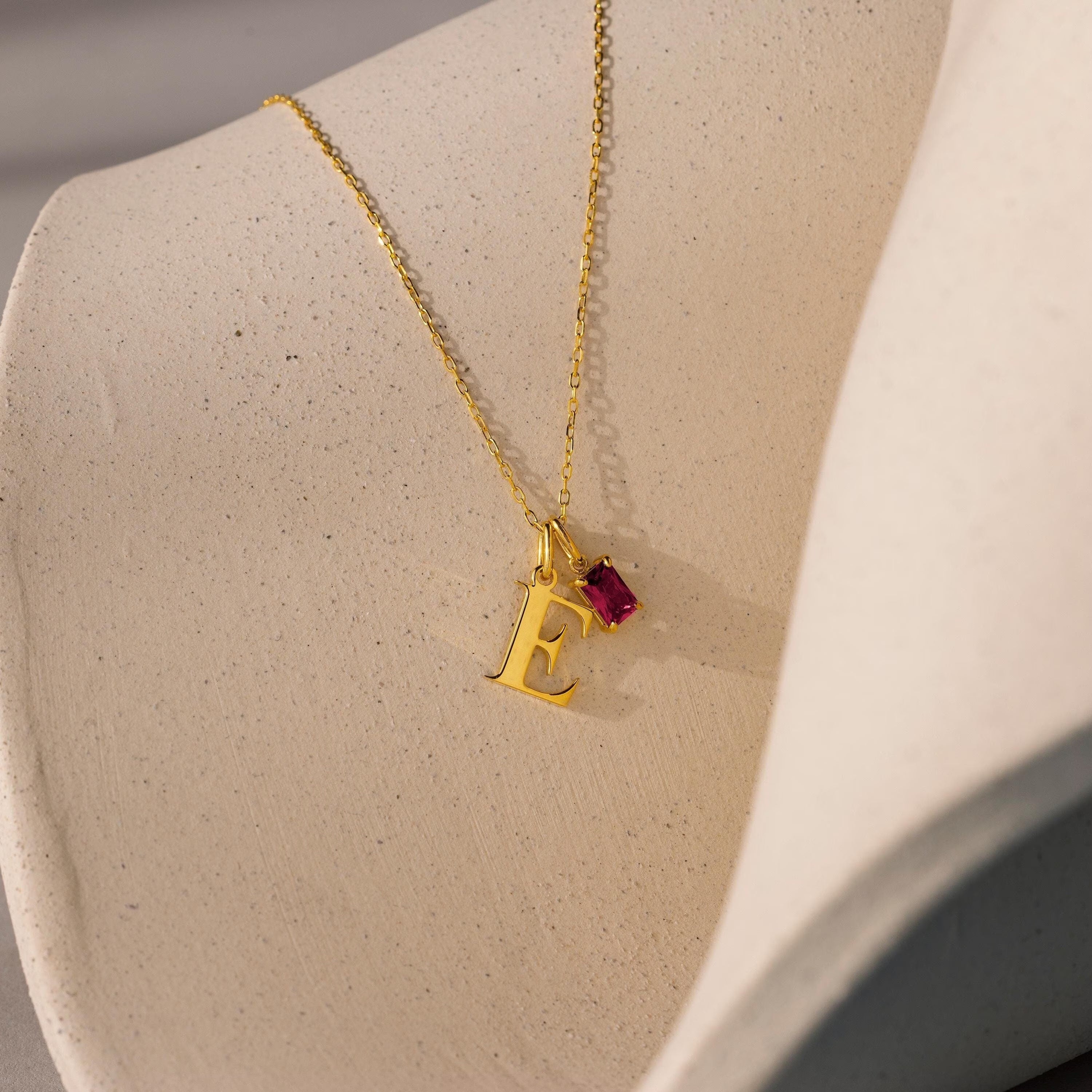 The Initial Birthstone Charm Necklace features a gold E pendant and a small red gemstone charm, beautifully showcased on a textured beige surface.
