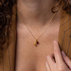 A person wearing the Initial Birthstone Charm Necklace, featuring a gold letter pendant and a small red gemstone charm, paired with a brown jacket.