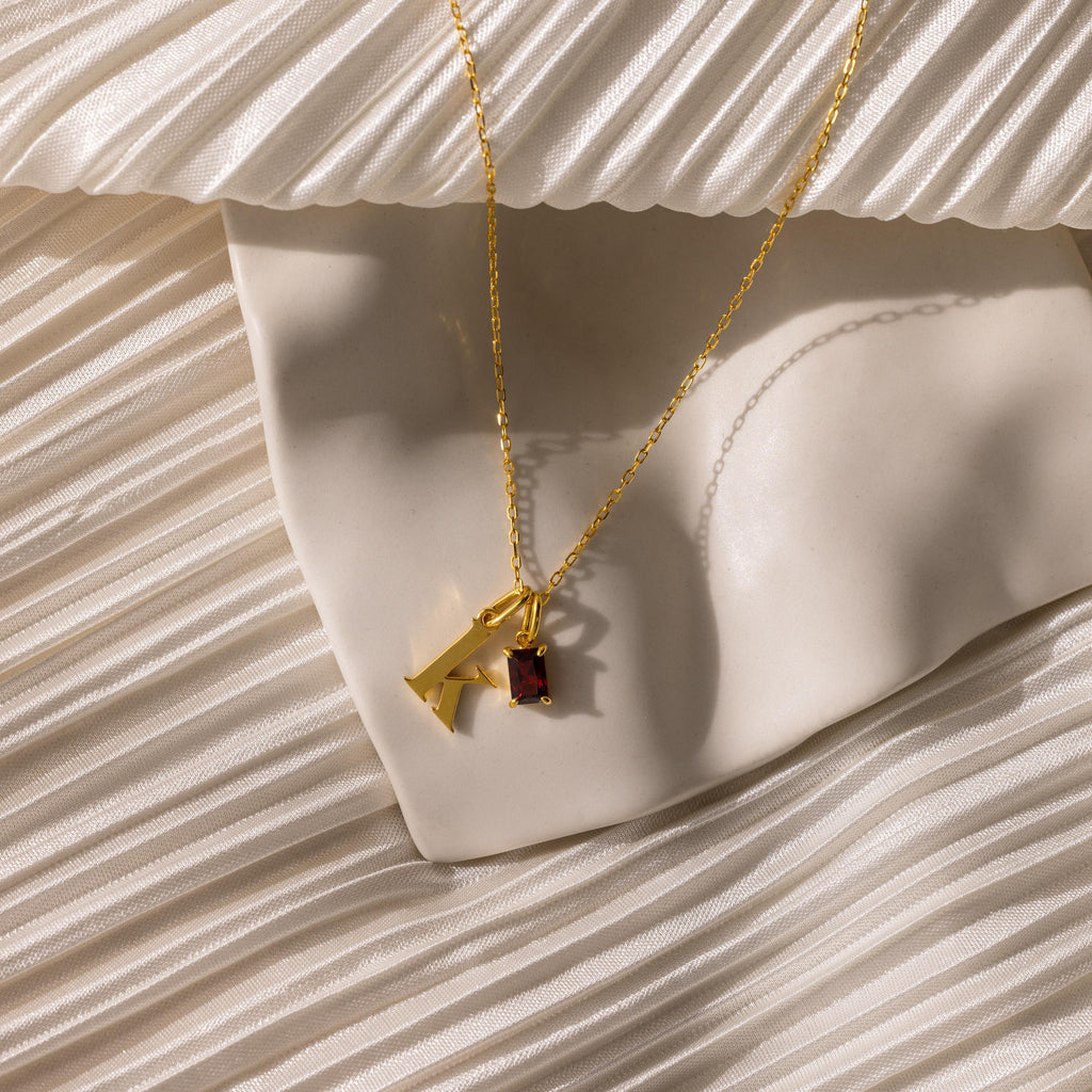 The Initial Birthstone Charm Necklace with a gold letter K and red gemstone pendant is shown on cream pleated fabric.
