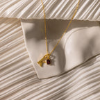 The Initial Birthstone Charm Necklace with a gold letter K and red gemstone pendant is shown on cream pleated fabric.