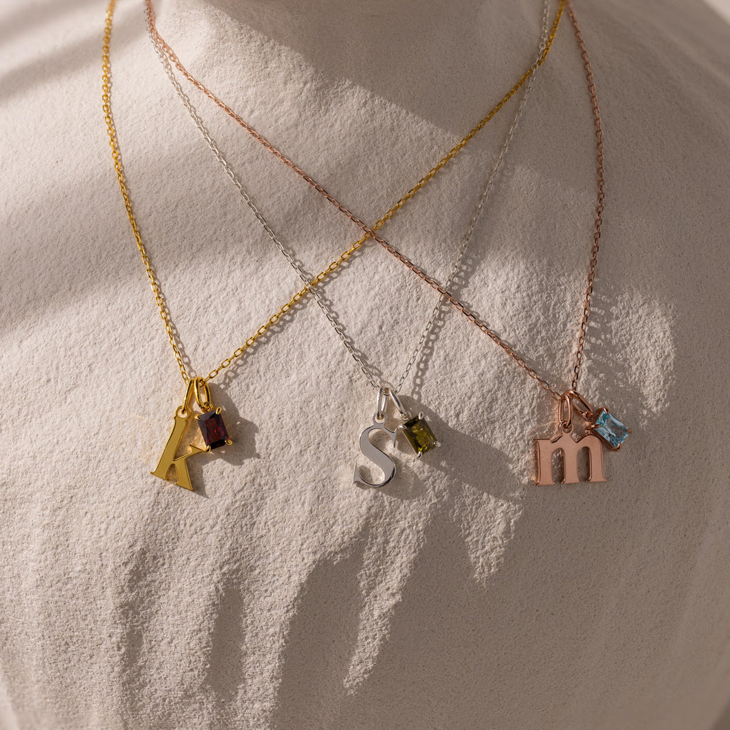 The Initial Birthstone Charm Necklace features three personalized pendants—A, S, and M—paired with vibrant gemstone charms, all displayed on a light textured surface.