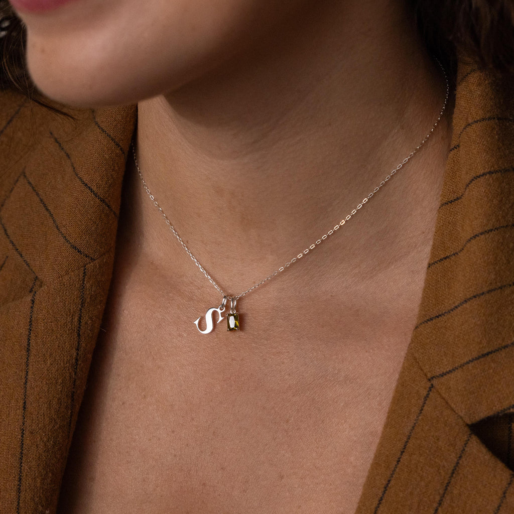 A woman wears the Initial Birthstone Charm Necklace, showcasing a silver “S” charm and small gemstone, styled with a brown pinstripe blazer.