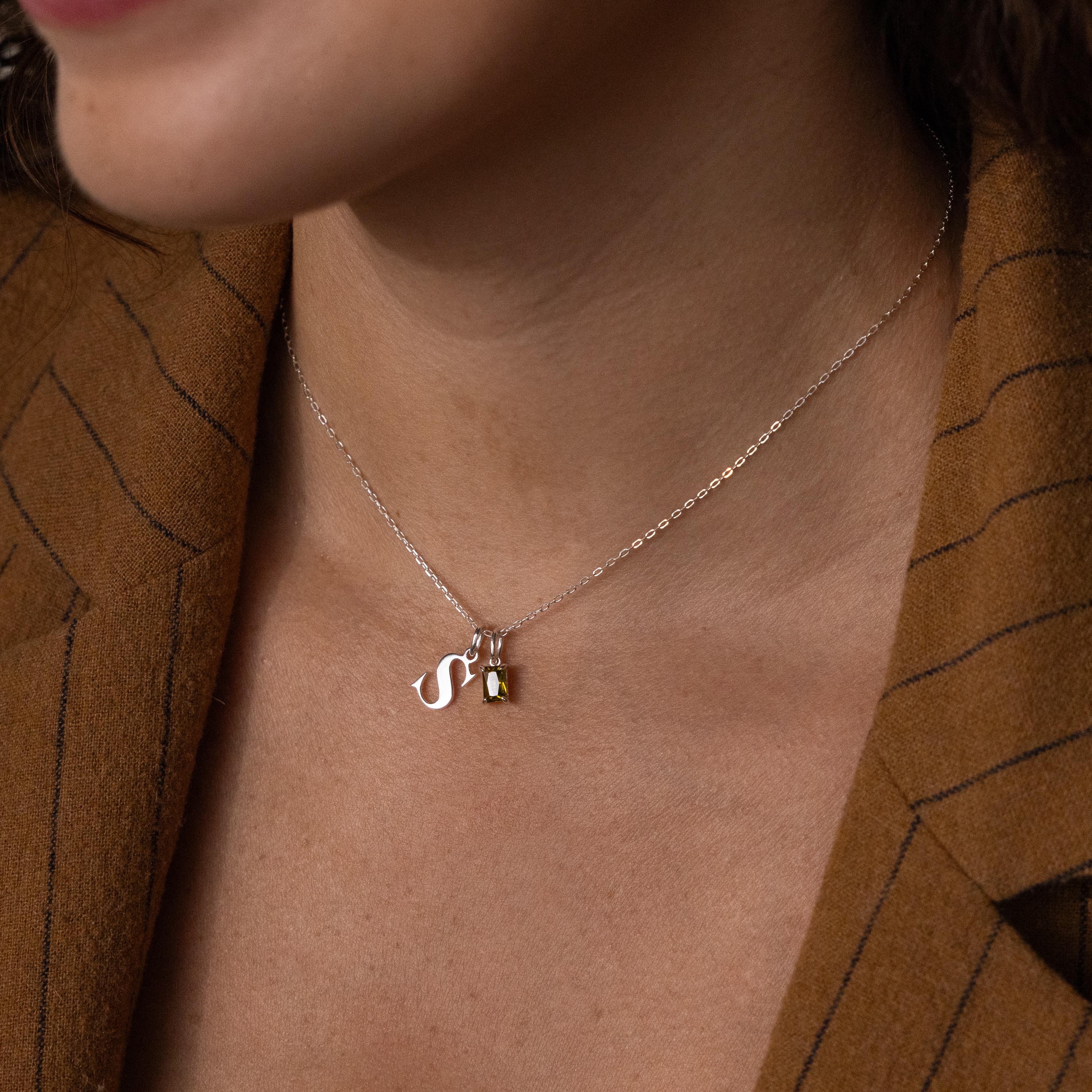 A woman wears the Initial Birthstone Charm Necklace, showcasing a silver “S” charm and small gemstone, styled with a brown pinstripe blazer.