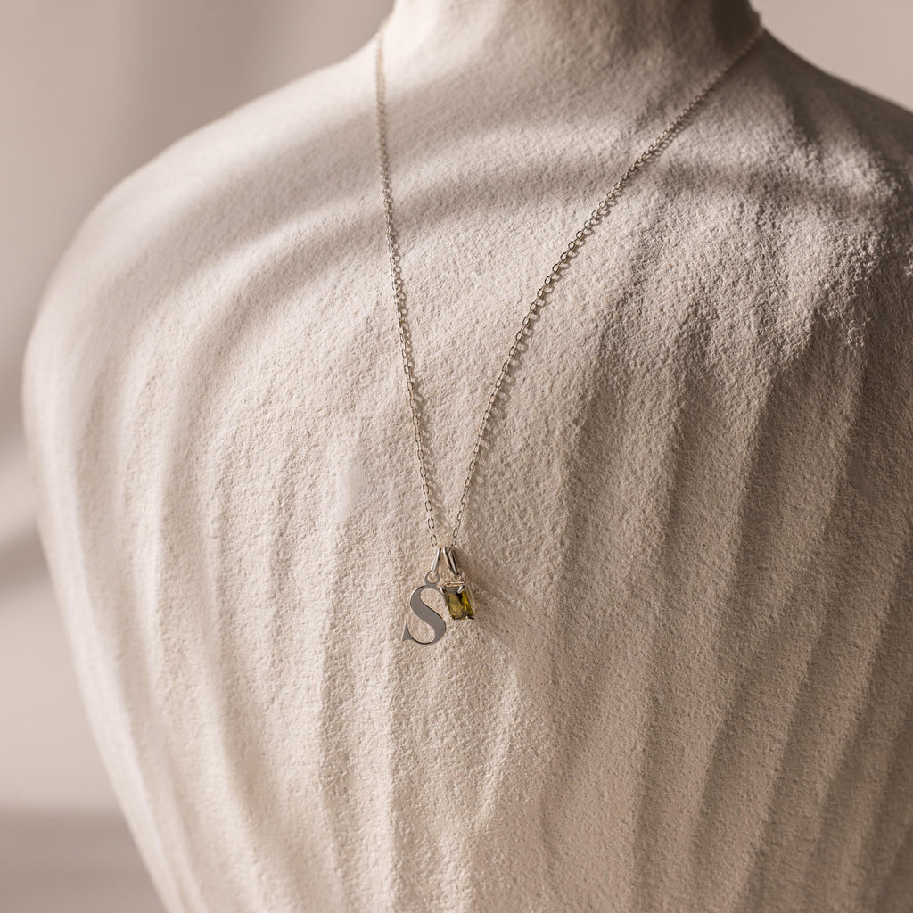 The Initial Birthstone Charm Necklace features a silver S pendant and a yellow gemstone charm, showcased on a textured white bust.