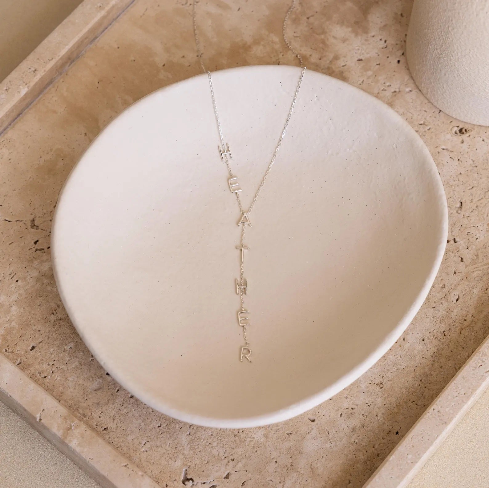 Lariat Name Necklace spelling HEATHER is displayed in a simple white ceramic bowl on a beige stone tray—a perfect piece of personalized jewelry.