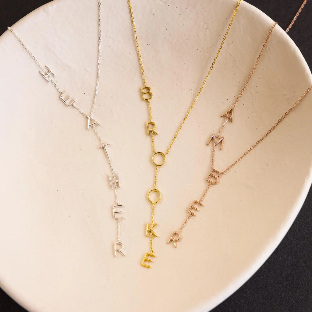 Three Lariat Name Necklaces with letter charms spelling “HIUAT THERE,” “BROOKE,” and “AMBER” are displayed on a white dish, highlighting unique personalized jewelry perfect for adding a special touch.