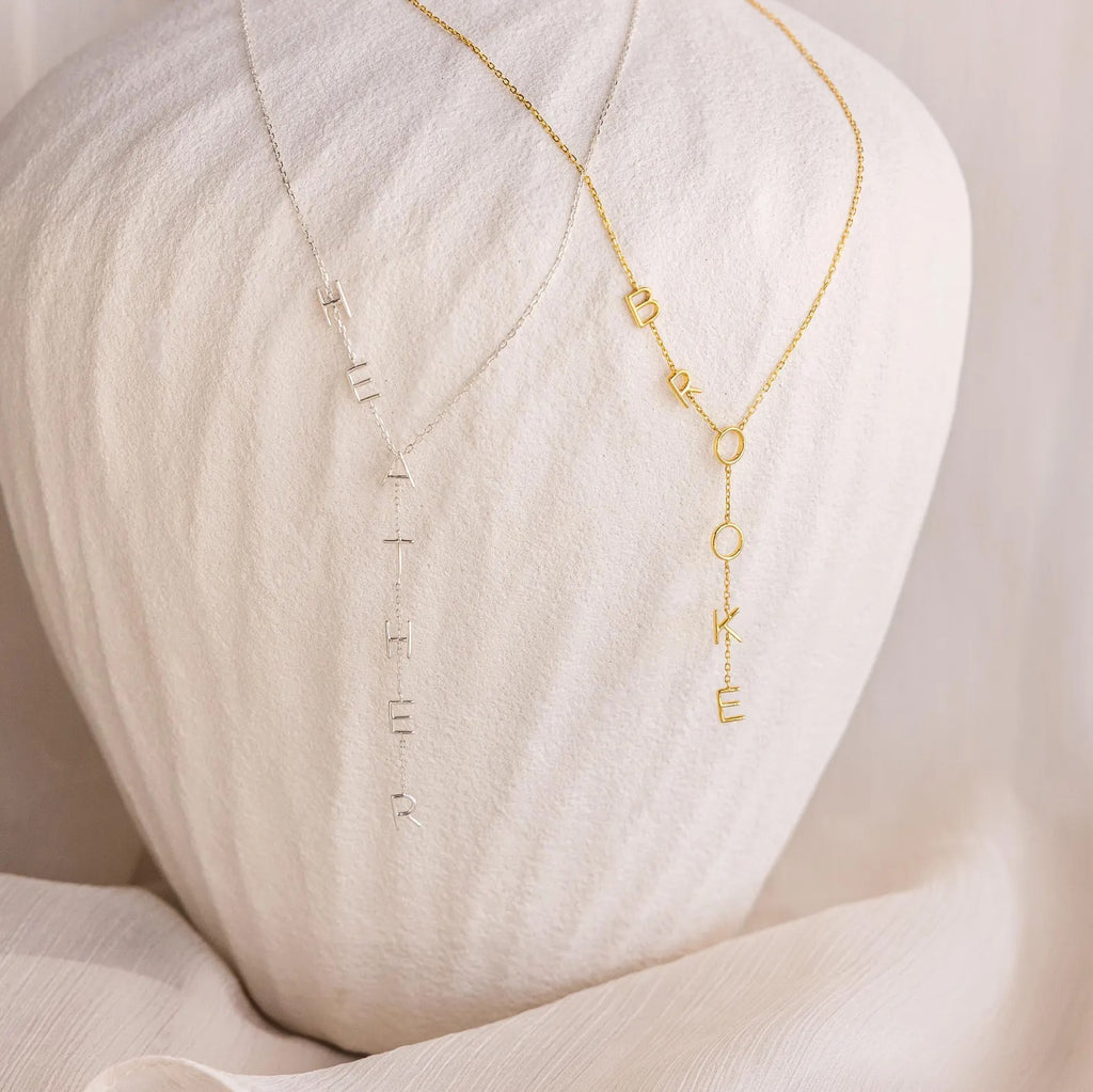 Two Lariat Name Necklaces with vertical letter charms spell HEATHER and BROOKE, displayed on a light textured bust to showcase elegant personalized jewelry.