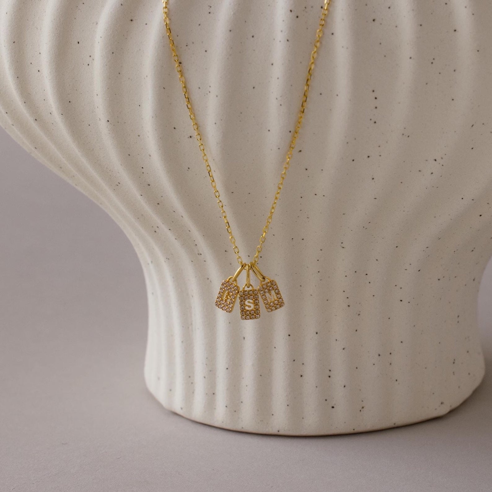 The Pave Initial Tag Necklace features three delicate charms and is shown on a textured white ceramic vase—a perfect personalized jewelry piece.