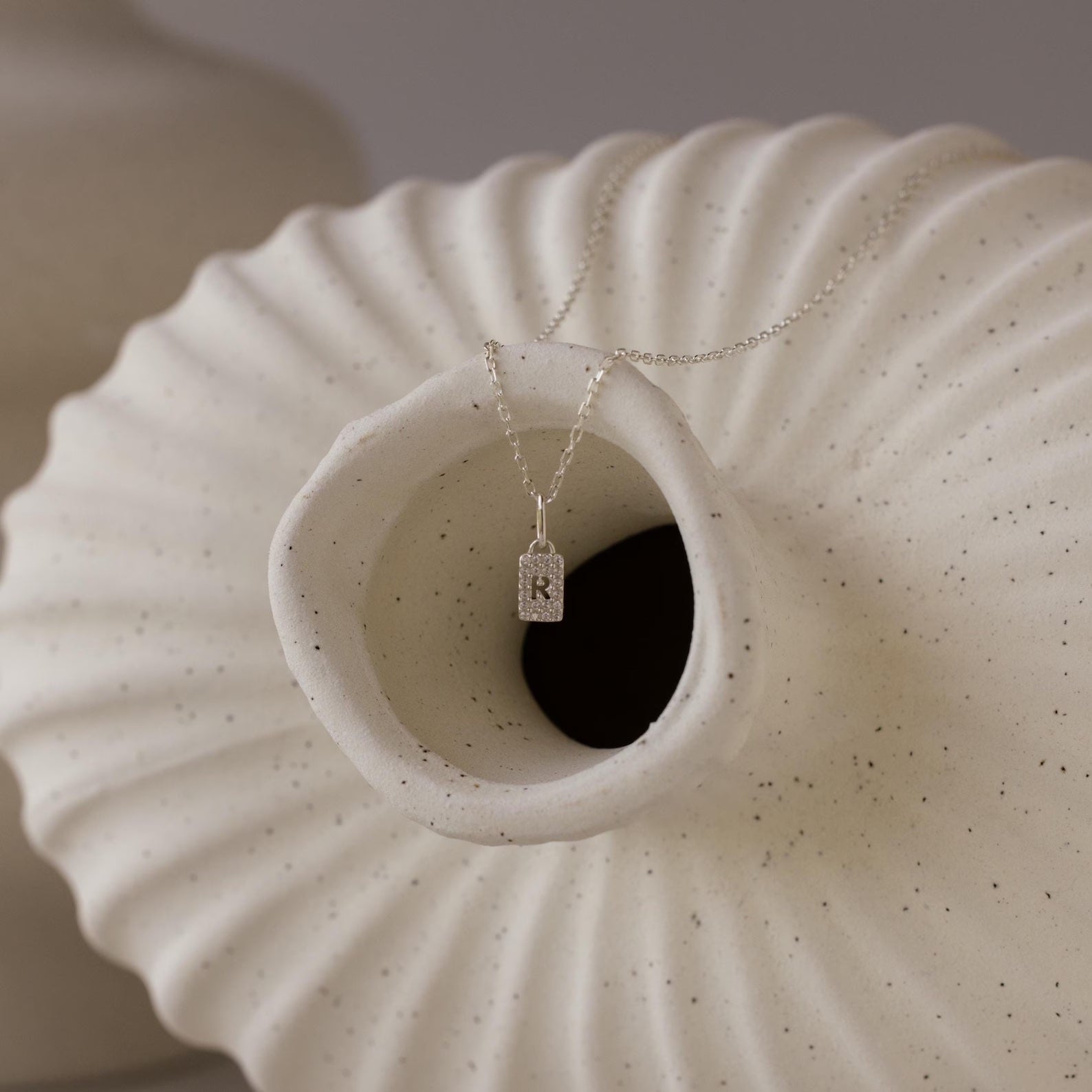 The Pave Initial Tag Necklace featuring an R pendant rests on the opening of a textured, off-white ceramic vase—an elegant and stylish piece of personalized jewelry.
