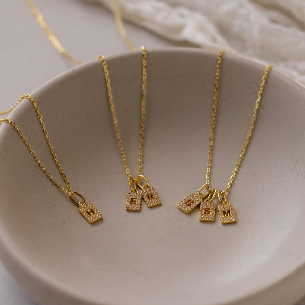 Discover the perfect personalized necklace for your collection with our Pave Initial Tag Necklace—featuring a gold chain and rectangular letter pendant, beautifully displayed on a beige ceramic dish. Elevate your initial jewelry style today!.