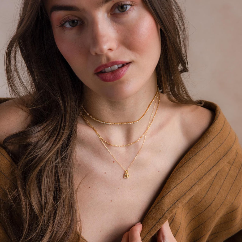 A woman with long brown hair wears a brown off-shoulder top and layered gold necklaces, featuring the Pave Initial Tag Necklace, as she looks at the camera.