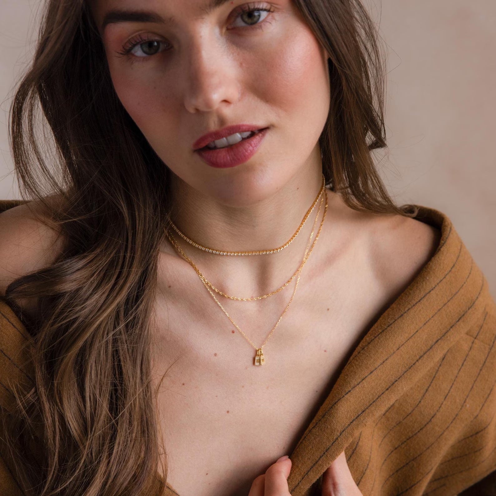 A woman with long brown hair wears a brown off-shoulder top and layered gold necklaces, featuring the Pave Initial Tag Necklace, as she looks at the camera.