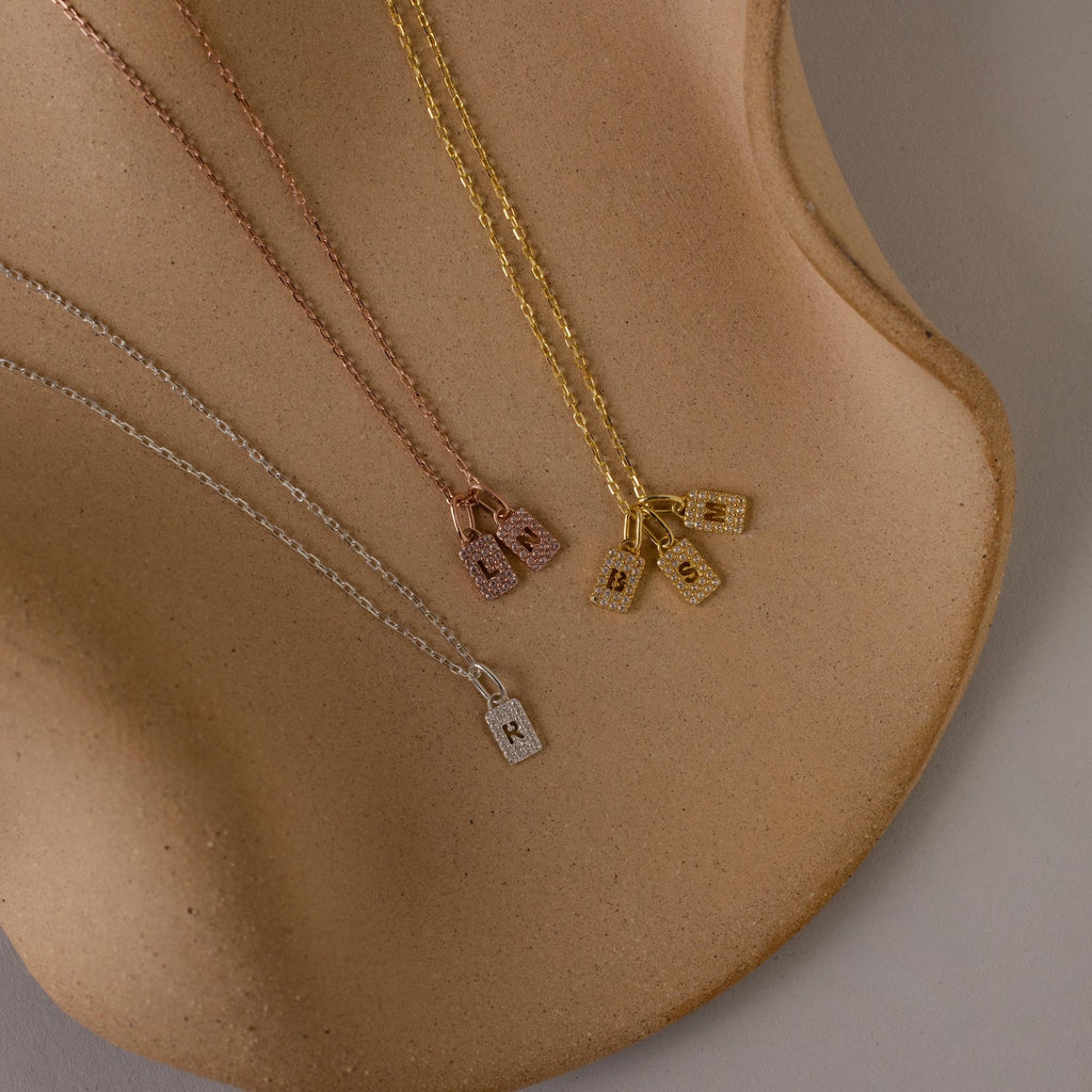 Three Pave Initial Tag Necklaces with lock pendants in gold, rose gold, and silver shown on a curved surface—ideal for stacking or adding to your initial jewelry collection.