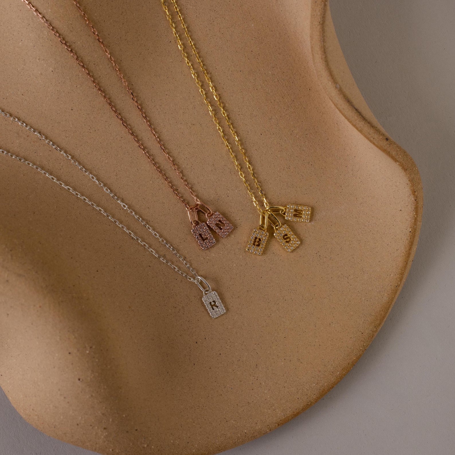 Three Pave Initial Tag Necklaces with lock pendants in gold, rose gold, and silver shown on a curved surface—ideal for stacking or adding to your initial jewelry collection.
