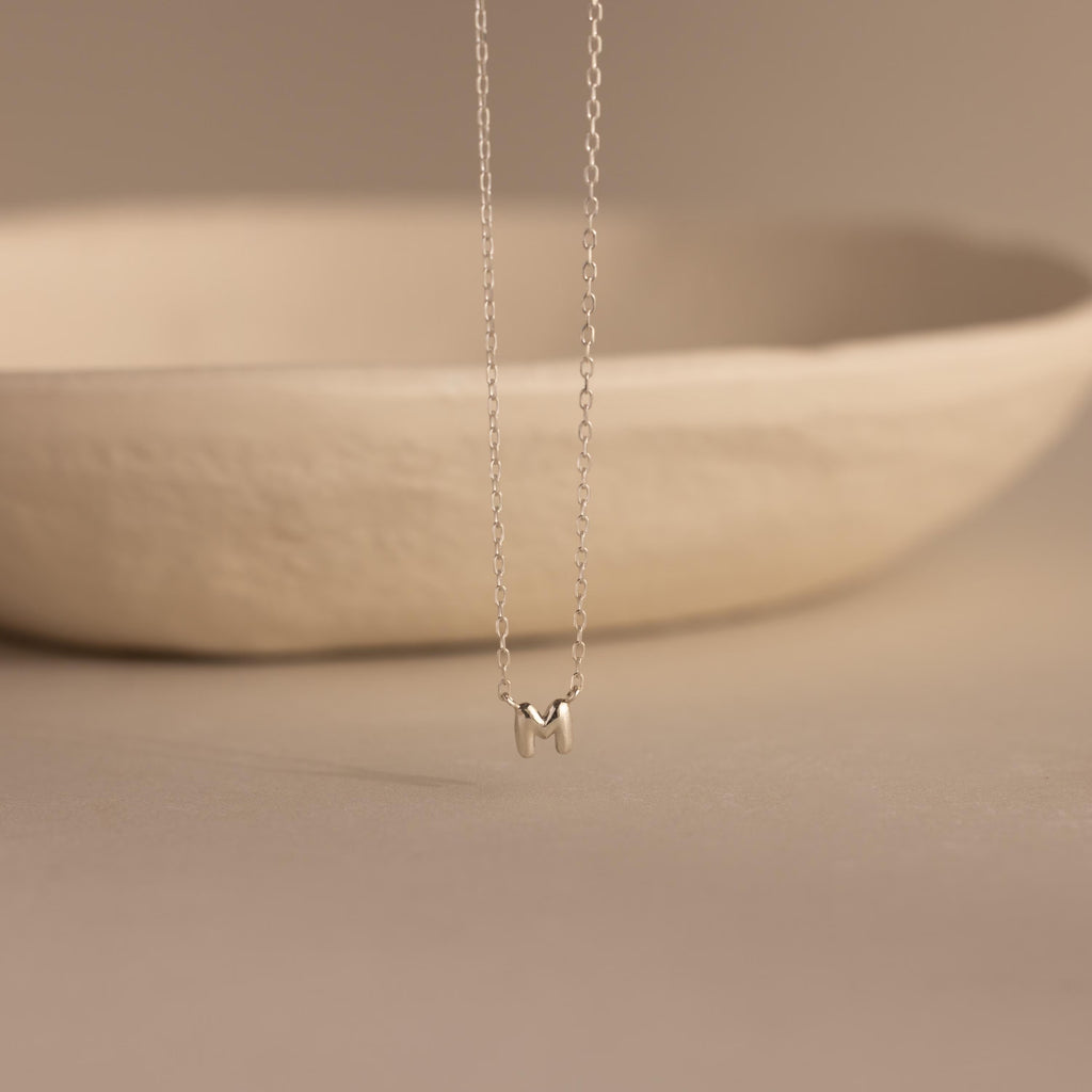 Silver dainty bubble initial M charm necklace against a neutral background.