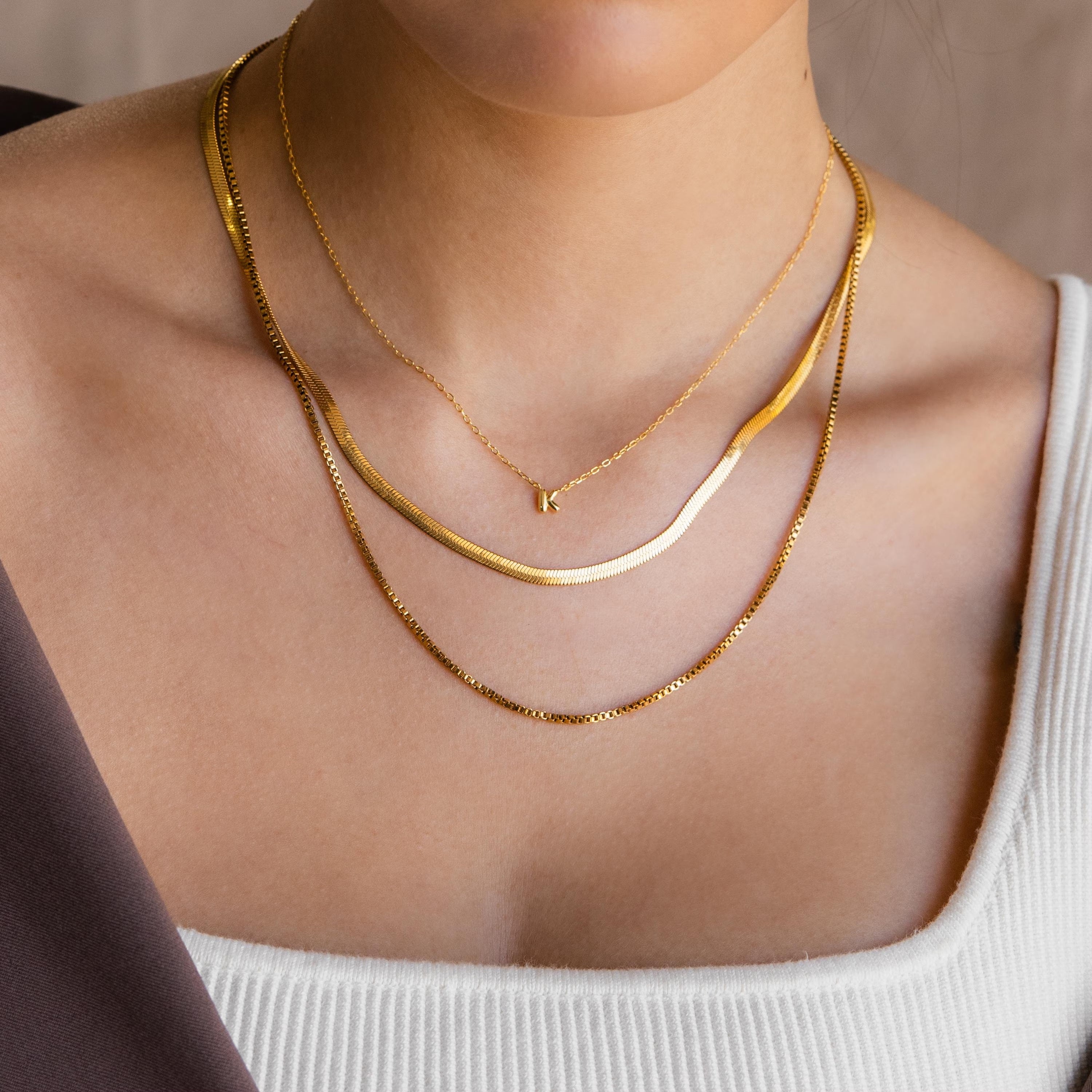 Model wearing a gold dainty bubble K letter charm necklace, layered with a gold herringbone necklace and gold box chain necklace.