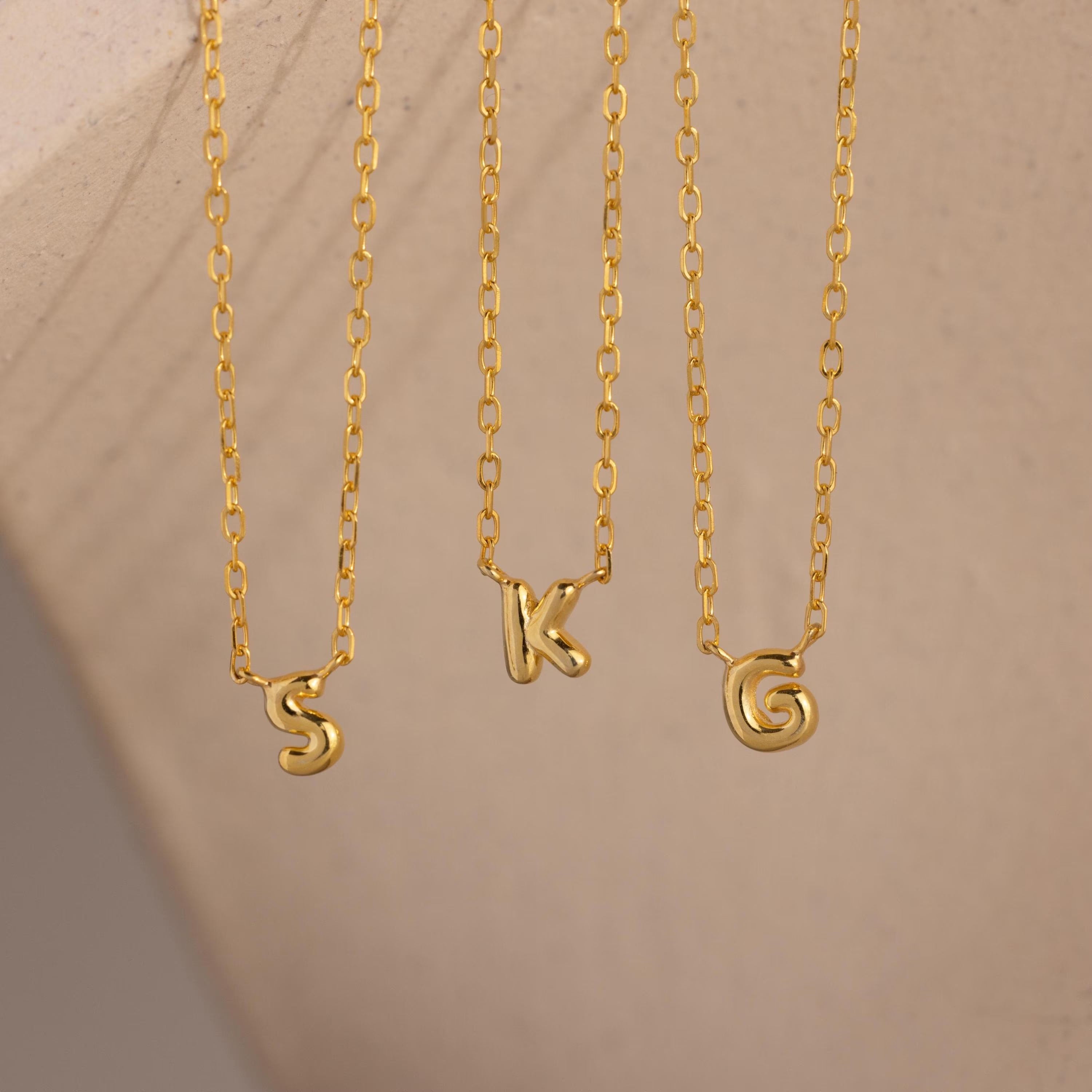 Three dainty gold Custom Bubble Initial Necklaces with the letters S, K, and G hanging side by side against a neutral background.