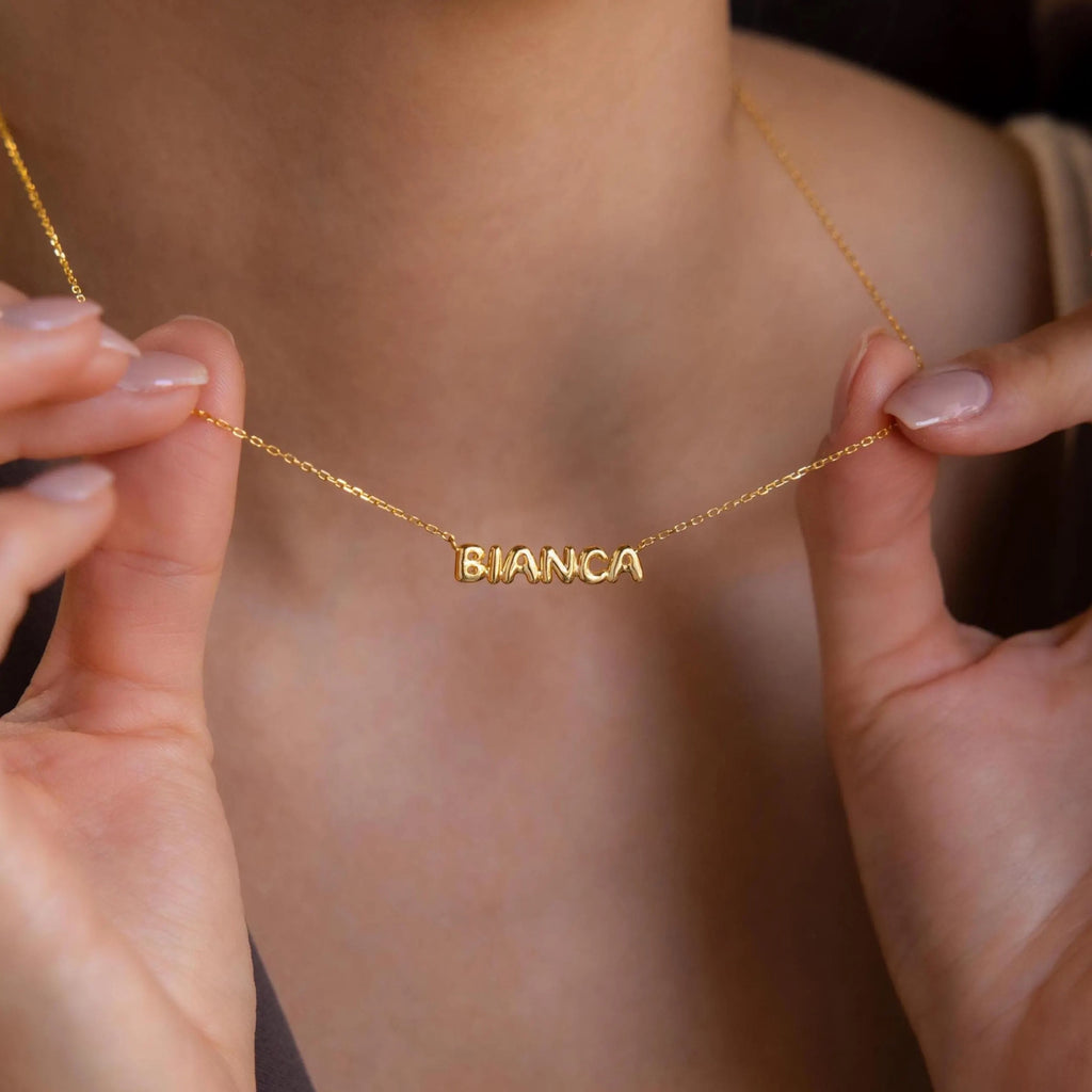 Model holding gold bubble name necklace with both hands, showcasing the word “BIANCA” in polished, chunky uppercase letters. The necklace has a cable chain and the letters have a smooth, slightly rounded design