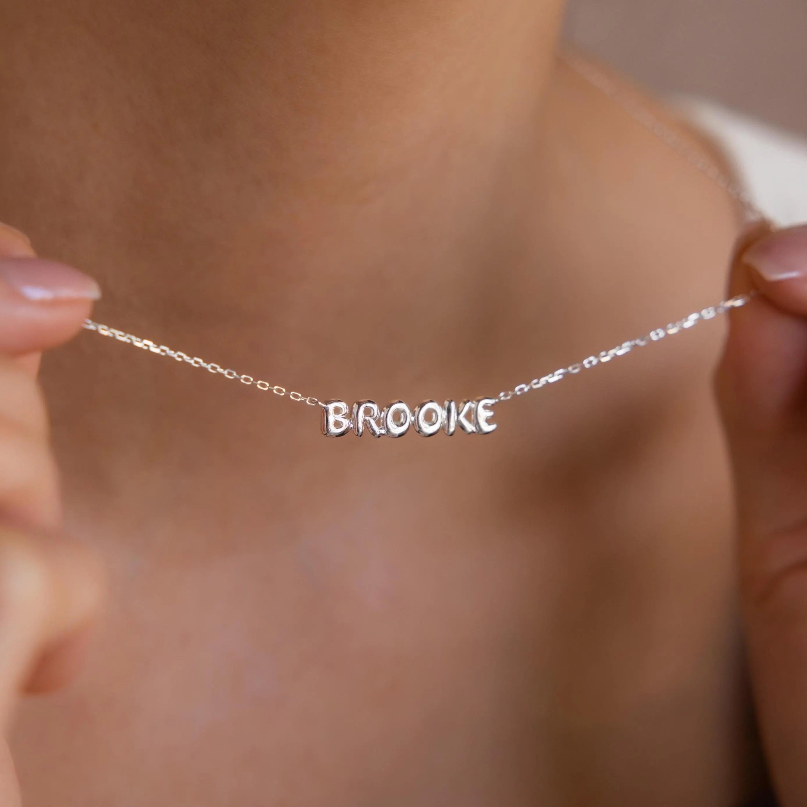 Model holds up silver bubble name necklace with name Brooke