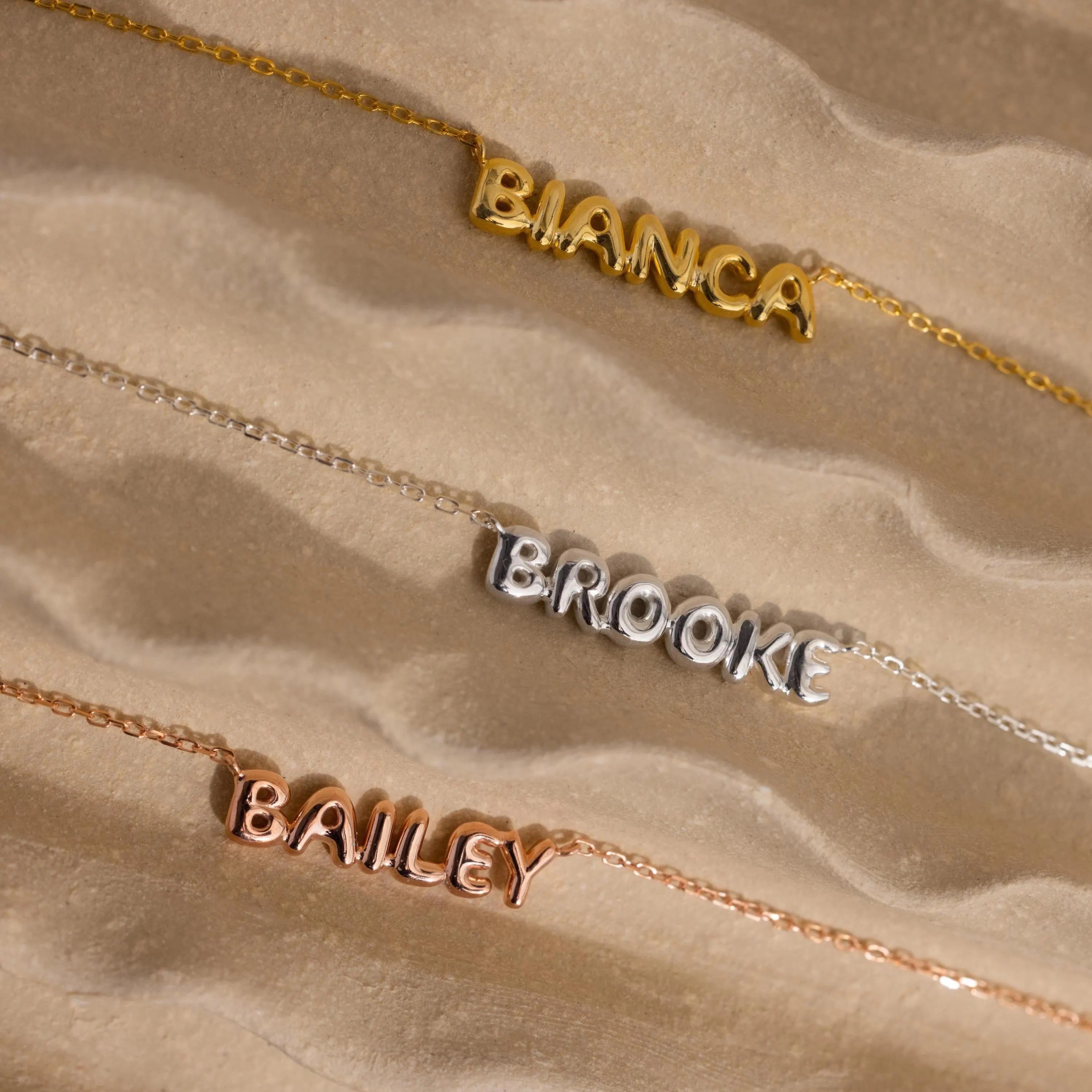 Gold Bianca bubble name necklace, silver Brooke necklace and rose gold Bailey necklace laying on sand.