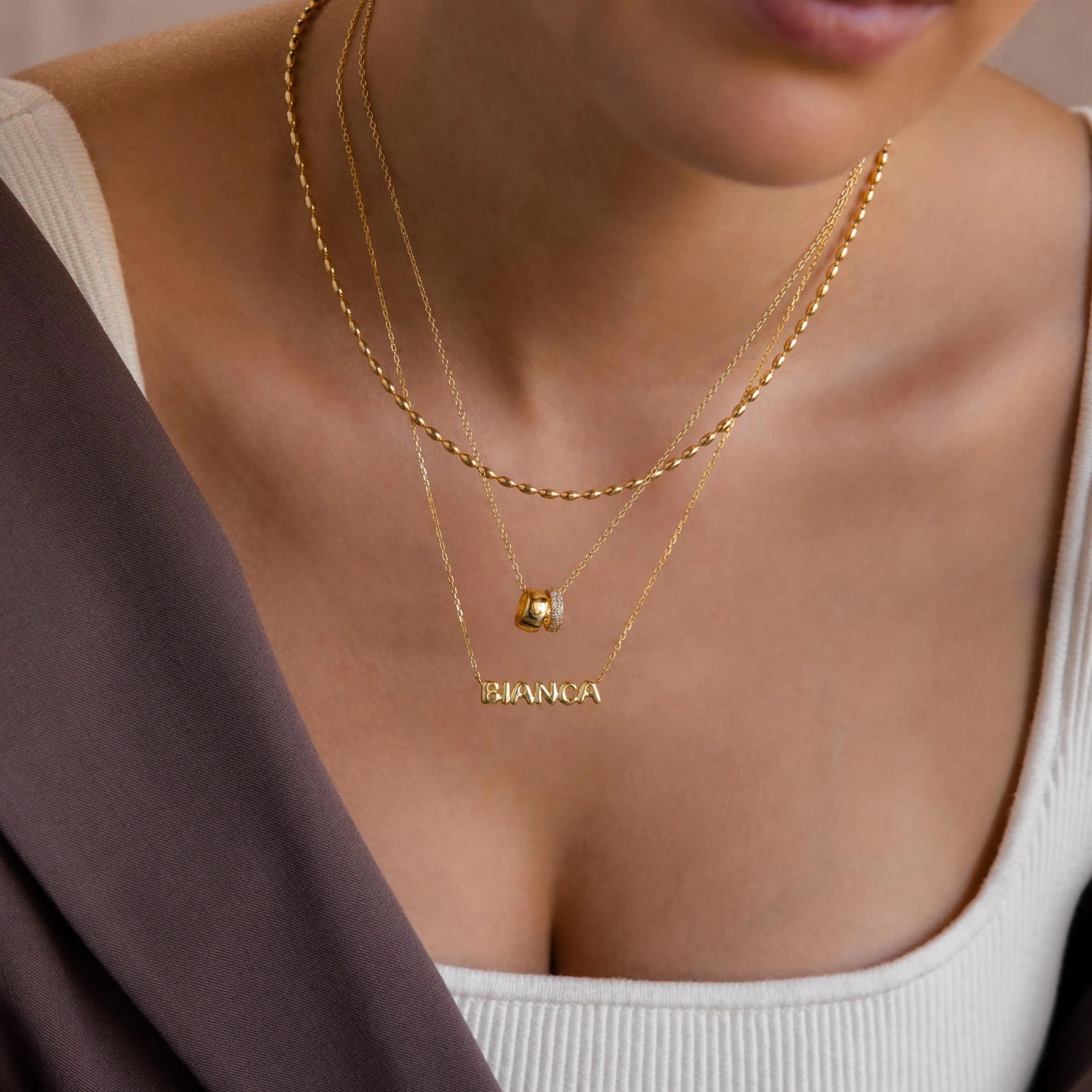 Model wears a gold bubble Bianca name necklace, layered with a pave duo necklace and oval beaded necklace on top.