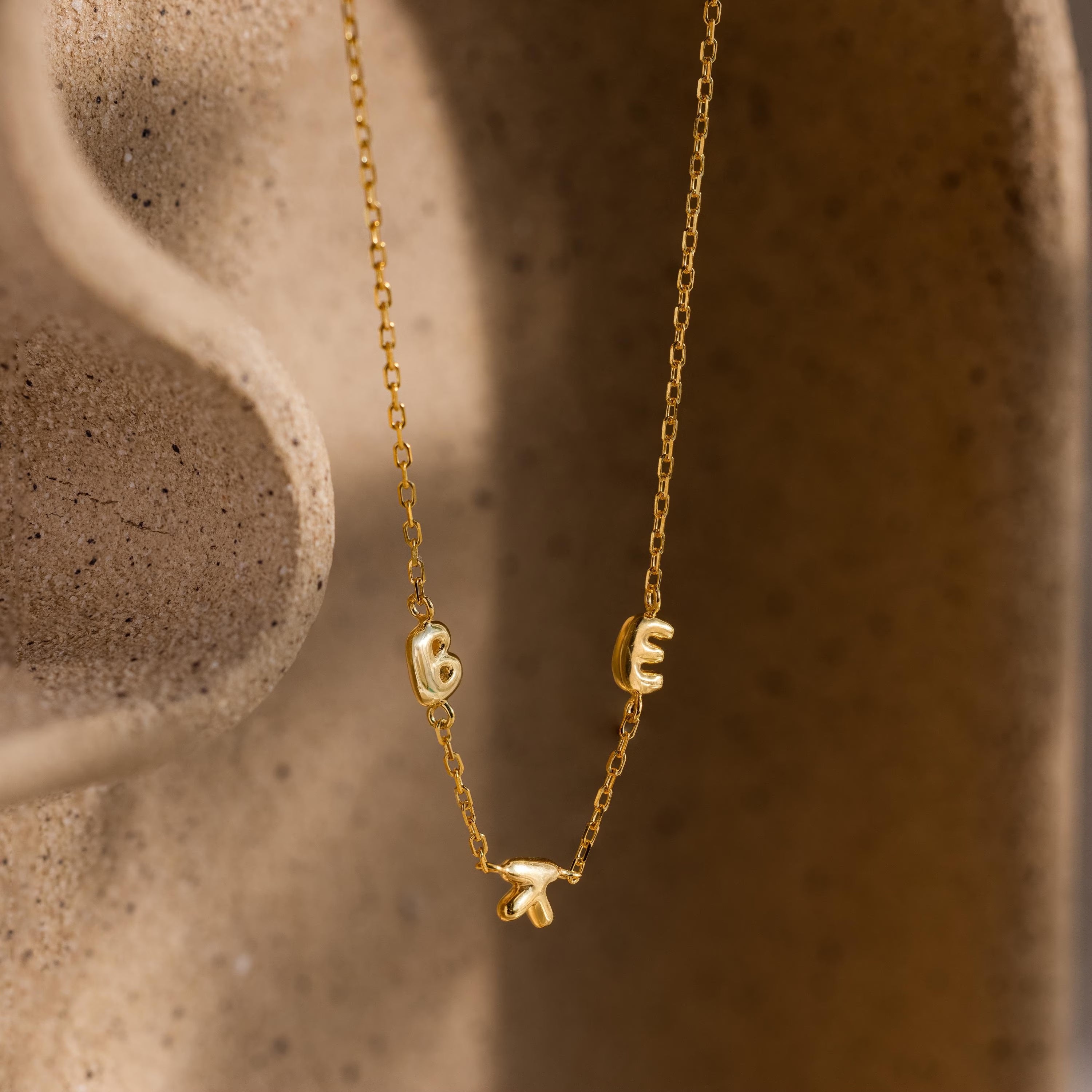Gold bubble letter necklace spelling “BEK” styled against a sculpted ceramic surface.