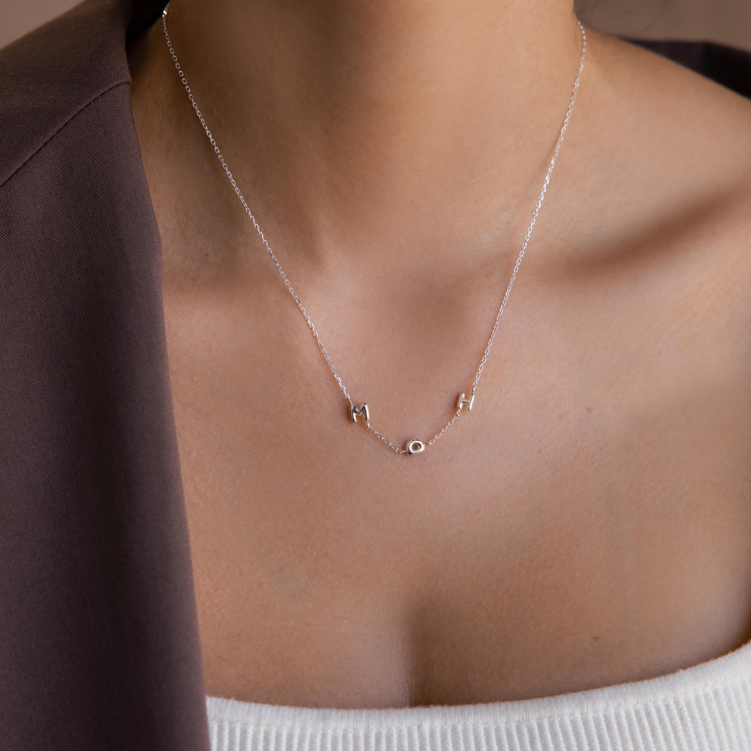 The Multiple Bubble Letter Necklace features silver bubble letters M and H, styled with a white top and brown jacket—a unique boho accessory that complements any outfit.