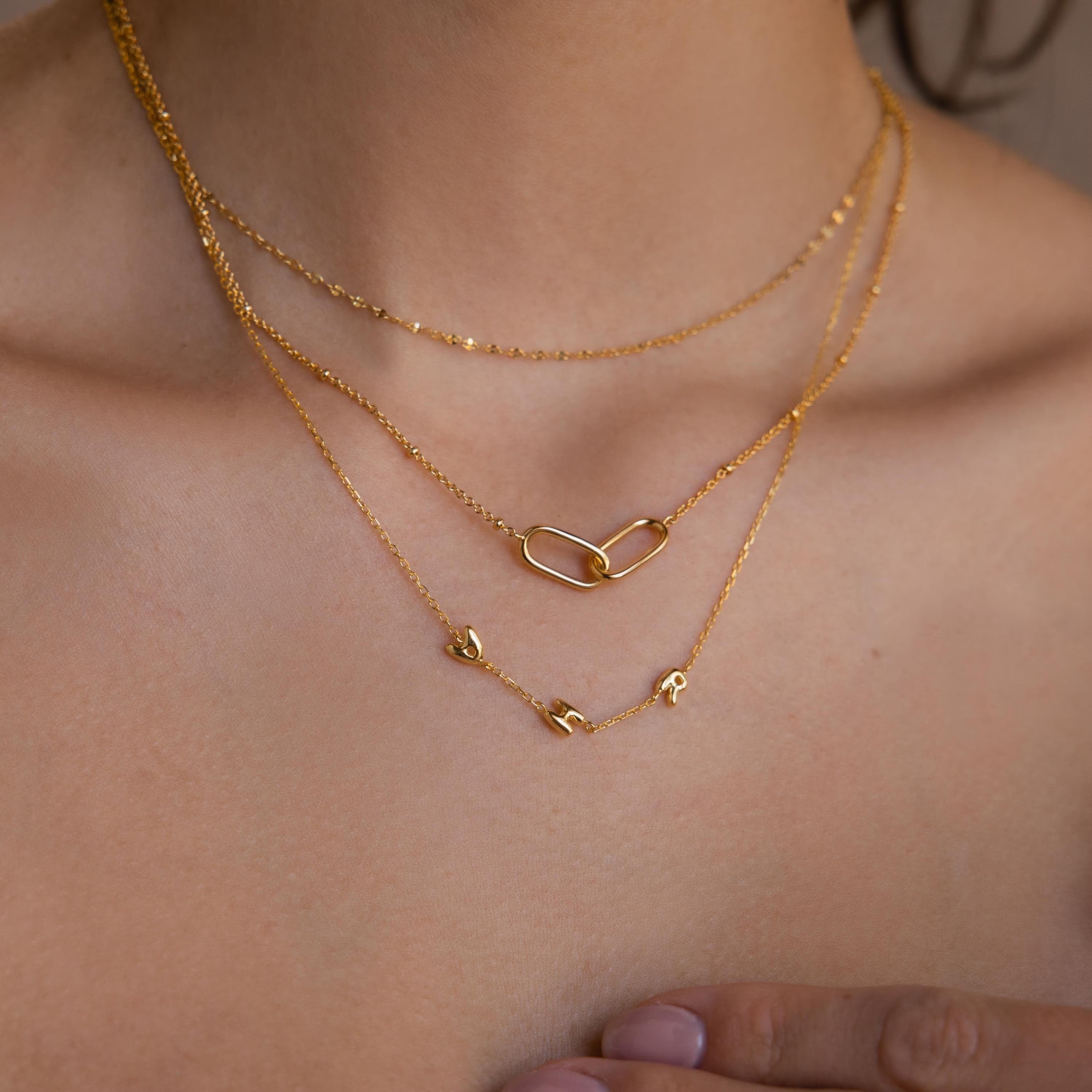 Model wears AHR gold bubble letter necklace, layered with gold linked pendant necklace and mirror chain.