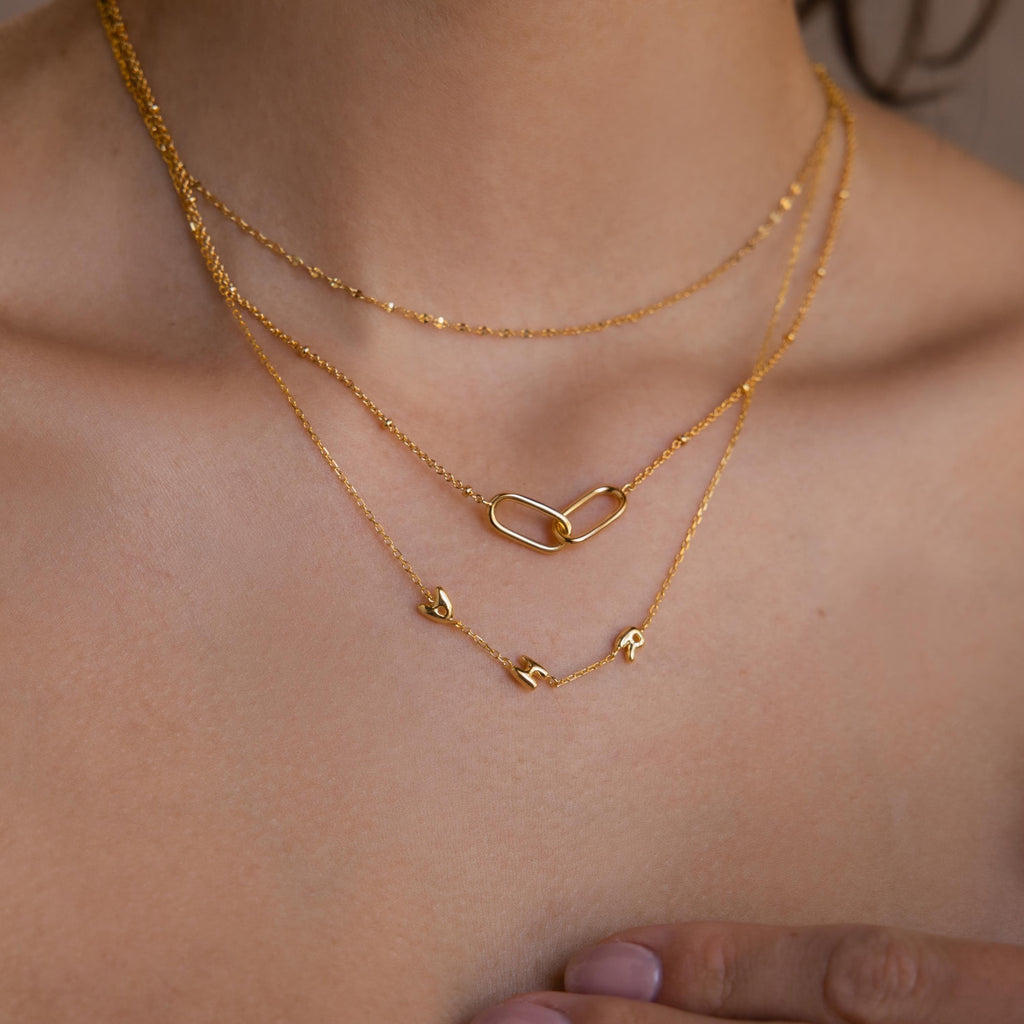 Model wearing a dainty gold bubble letter necklace, layered with a linked pendant necklace and mirror chain necklace.