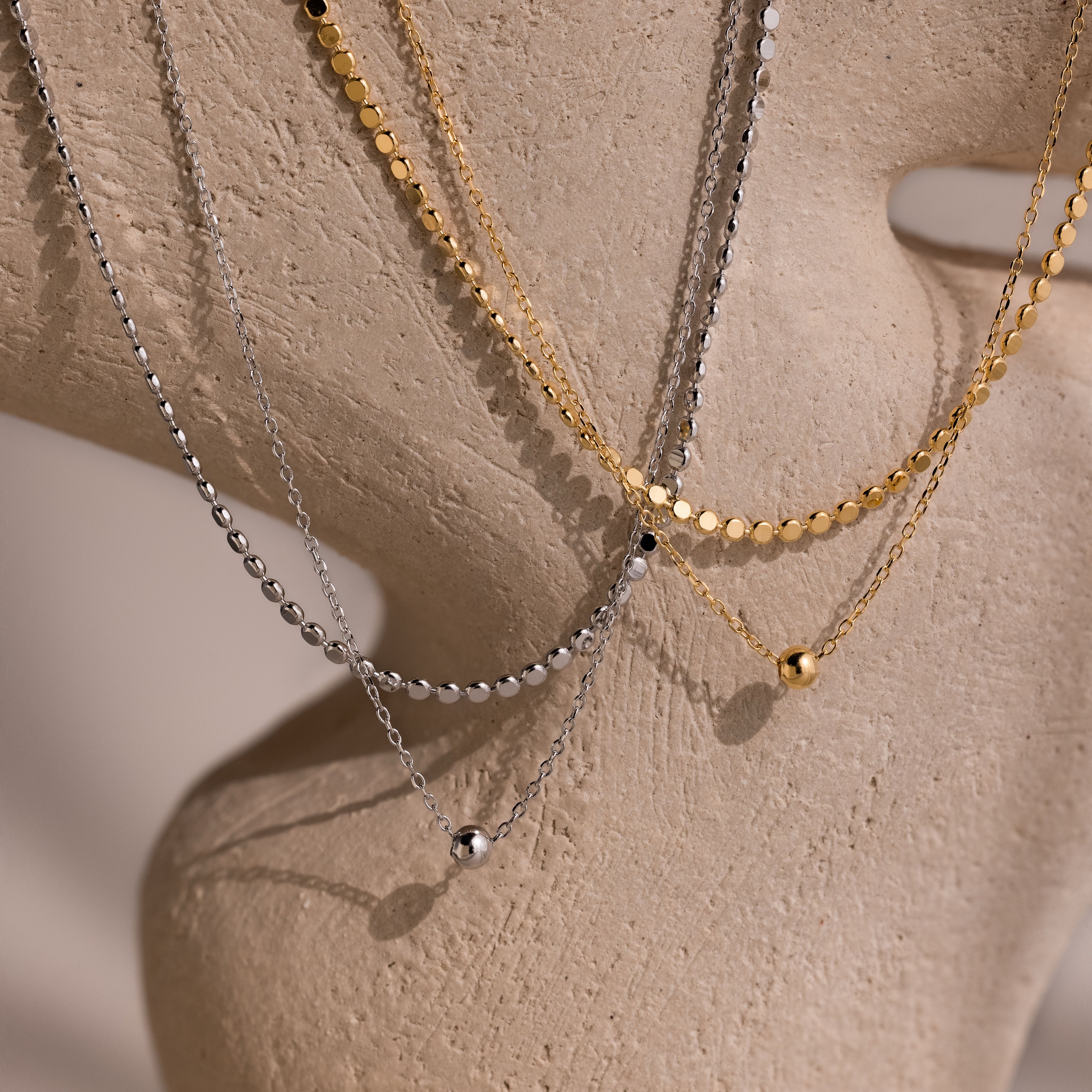 Two gold and silver beaded duo chain necklaces on a beige stone background.