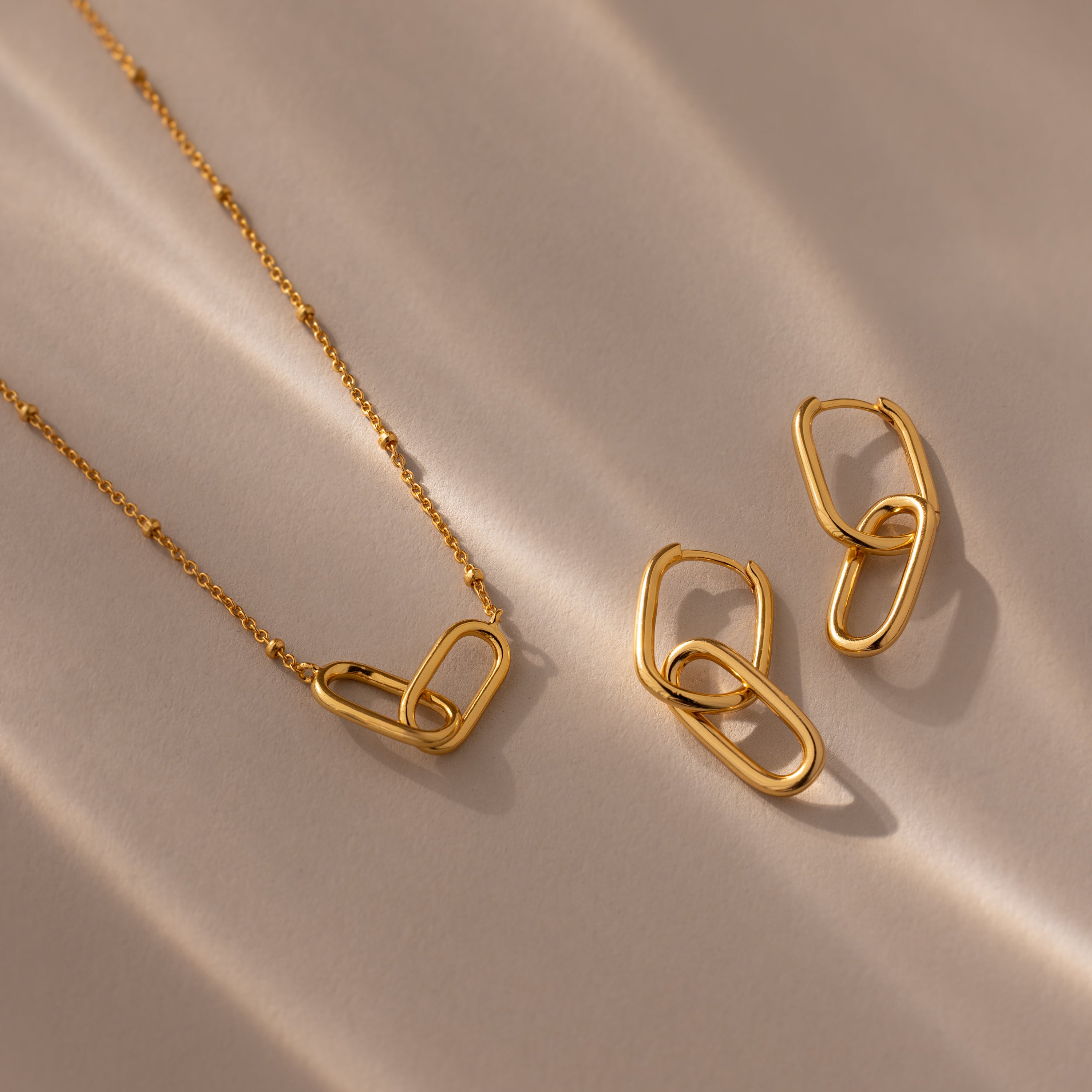 The Linked Charm Necklace and Earrings Set is displayed on a beige surface with soft light and shadows—a timeless anniversary gift for someone special.
