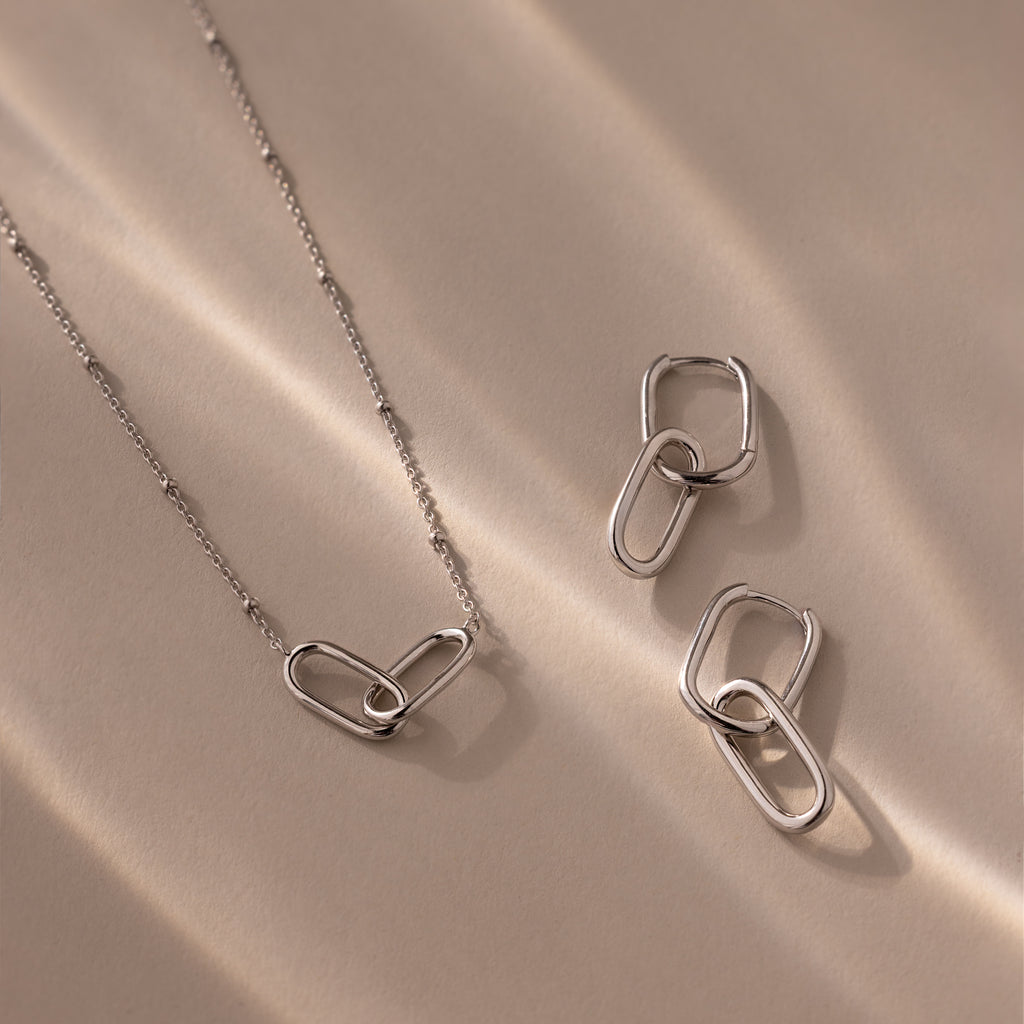 The Linked Charm Necklace and Earrings Set in Sterling Silver features interlocking oval links, elegantly displayed on a beige surface with soft shadows—an ideal anniversary gift.