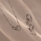 The Linked Charm Necklace and Earrings Set in Sterling Silver features interlocking oval links, elegantly displayed on a beige surface with soft shadows—an ideal anniversary gift.