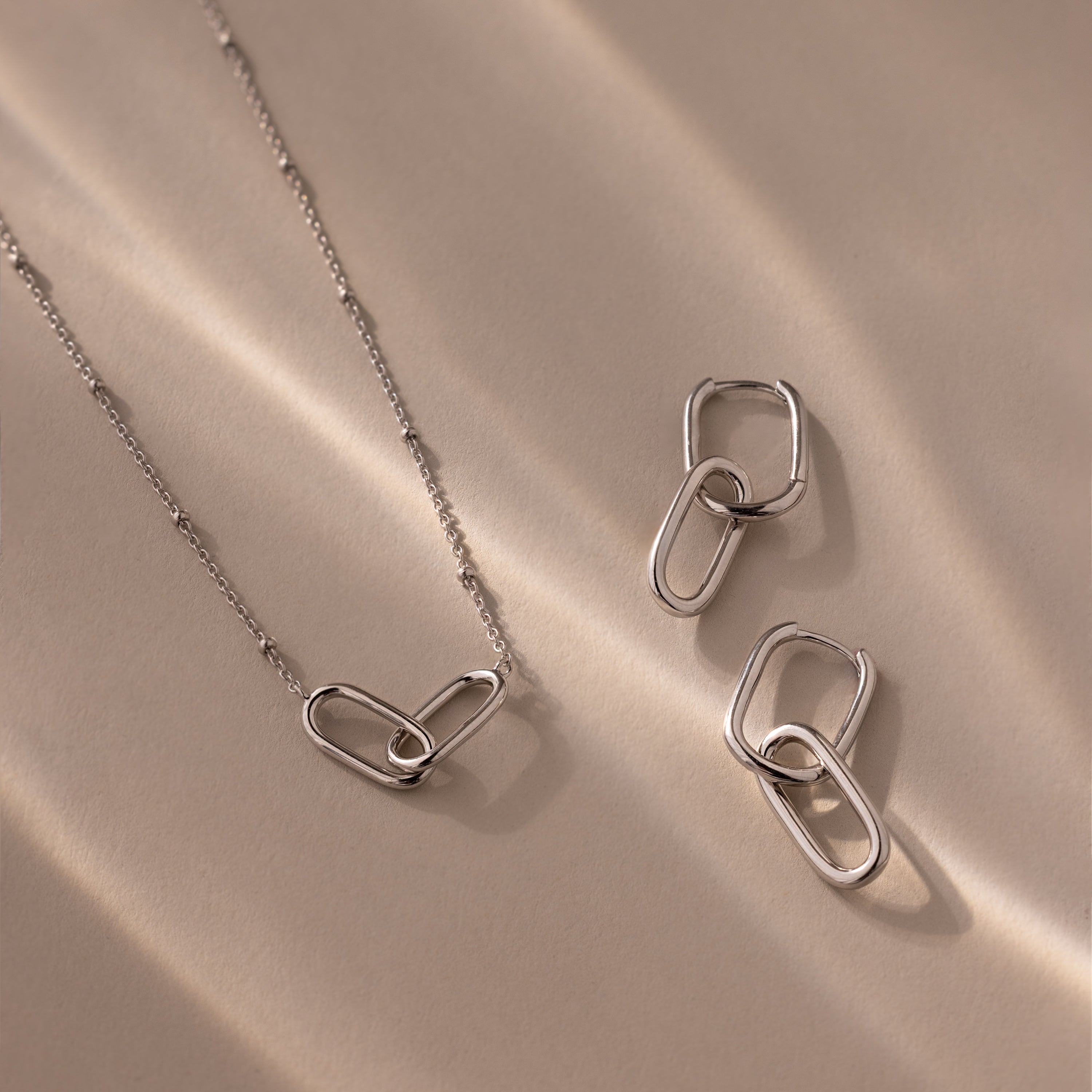 The Linked Charm Necklace and Earrings Set in Sterling Silver features interlocking oval links, elegantly displayed on a beige surface with soft shadows—an ideal anniversary gift.