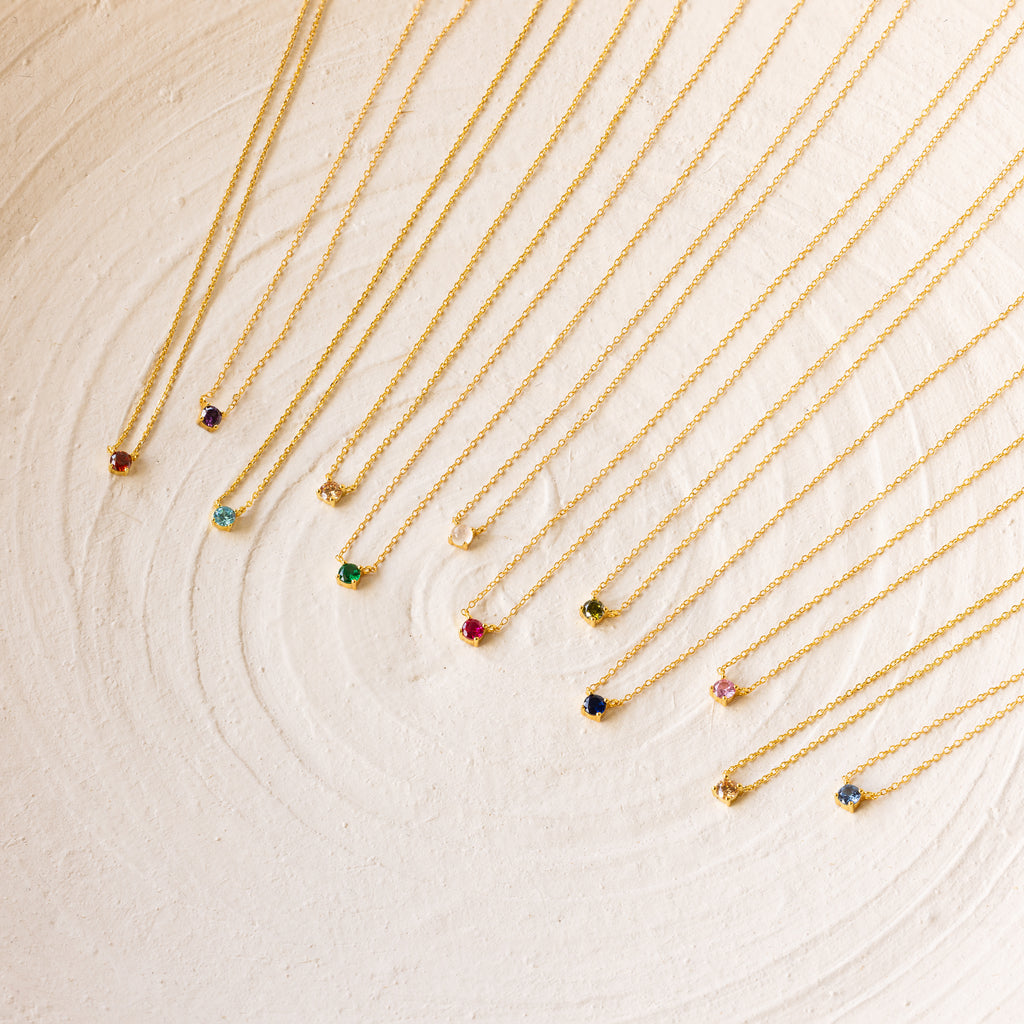 Twelve Dainty Birthstone Necklaces with colorful gemstone pendants are arranged in a neat row on a textured white surface, perfect for those seeking meaningful jewelry.