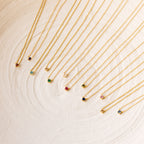 Twelve Dainty Birthstone Necklaces with colorful gemstone pendants are arranged in a neat row on a textured white surface, perfect for those seeking meaningful jewelry.