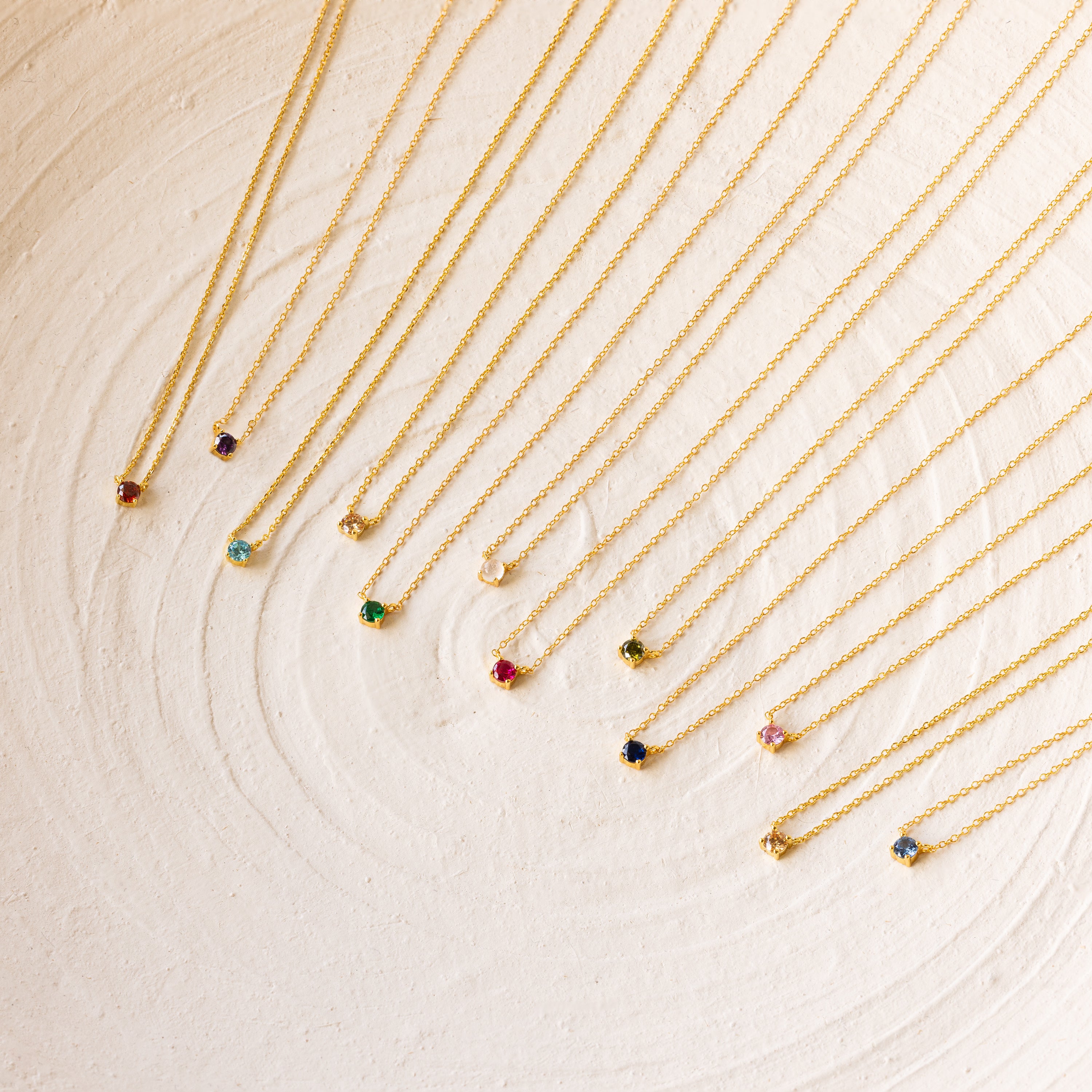 Twelve Dainty Birthstone Necklaces with colorful gemstone pendants are arranged in a neat row on a textured white surface, perfect for those seeking meaningful jewelry.
