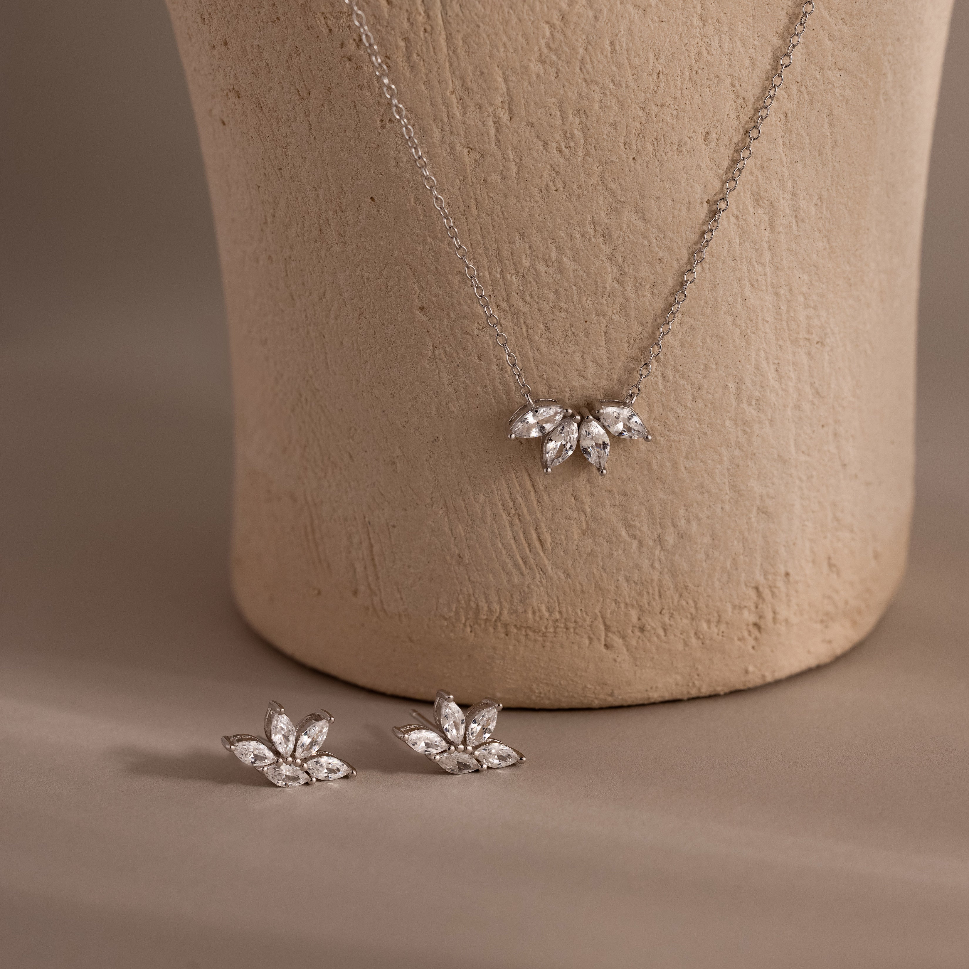 The Marquise Diamond Jewelry Set in Sterling Silver features a silver necklace and matching earrings with leaf designs, showcased against a textured beige vase.