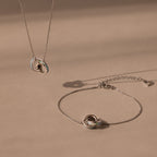 The Interlocking Opal Inlay Necklace and Bracelet Set in Silver features a knot pendant necklace with a matching bracelet, beautifully displayed against a neutral beige background.